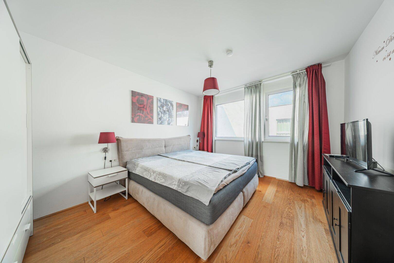 Gemütliches Schlafzimmer mit Doppelbett, Holzboden und roten Vorhängen, gut beleuchtet.