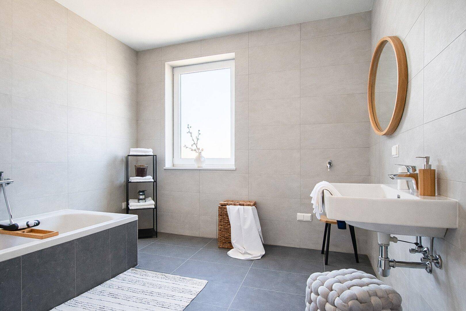 Modernes Badezimmer mit hellgrauen Fliesen, Badewanne und Fenster für Tageslicht.