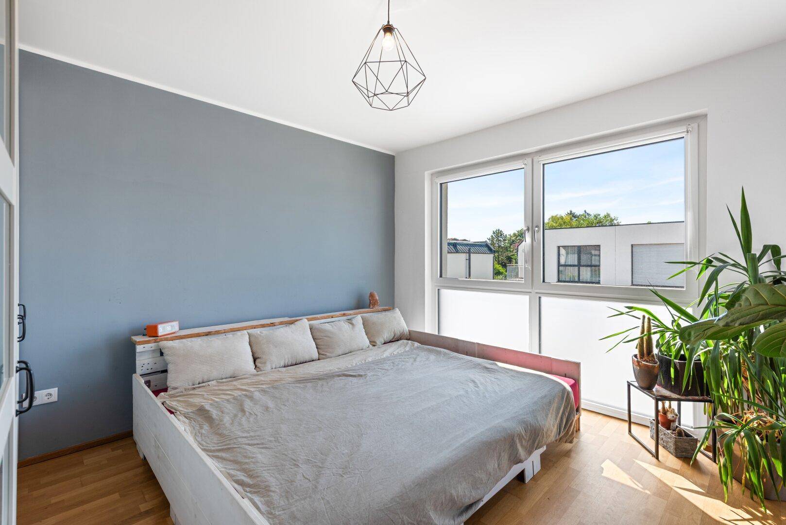 Gemütliches Schlafzimmer mit Holzboden, grauer Akzentwand und großem Fenster für viel Licht.