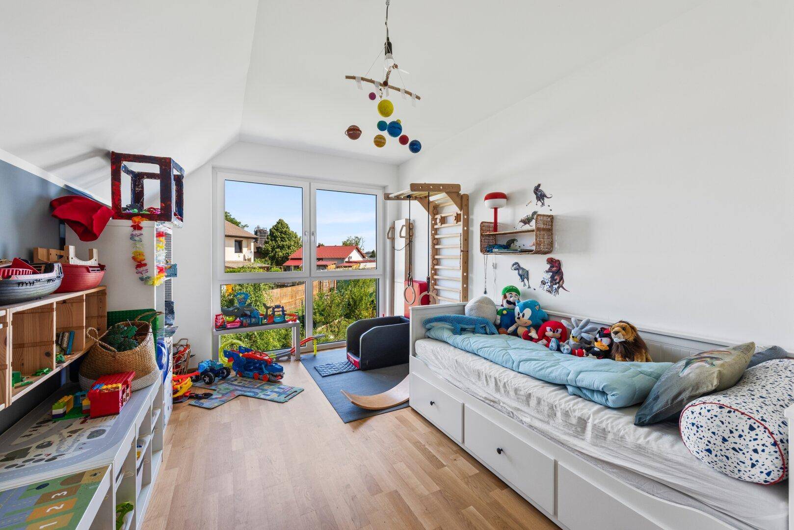 Helles Kinderzimmer mit Spielzeug, Holzboden und Fenster mit Blick ins Grüne.