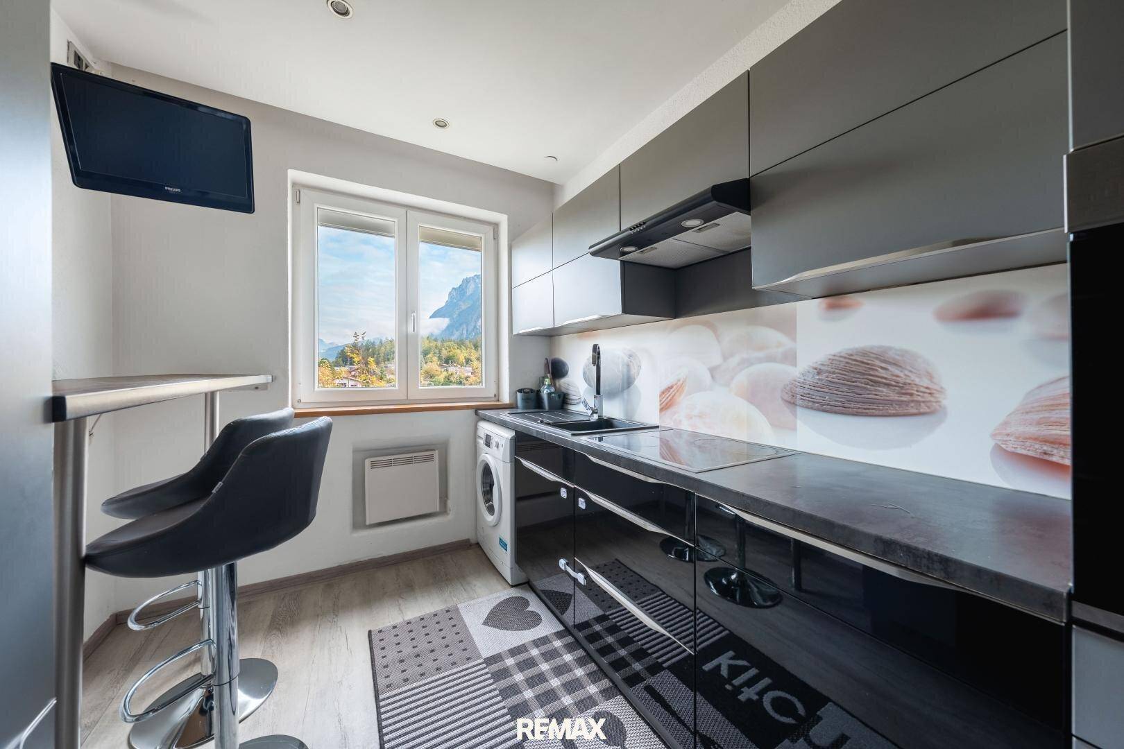 Funktionale Küche mit Fenster, Barhocker, Waschmaschine und Blick auf die Berge.