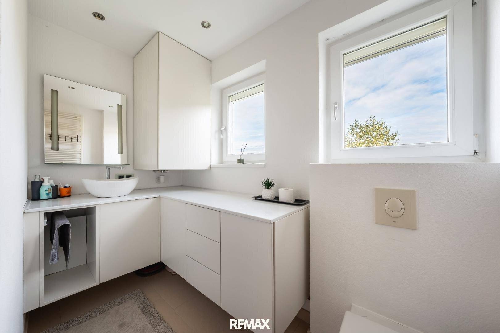 Modernes Badezimmer mit weißem Waschtisch, Unterschränken und zwei Fenstern für Tageslicht.