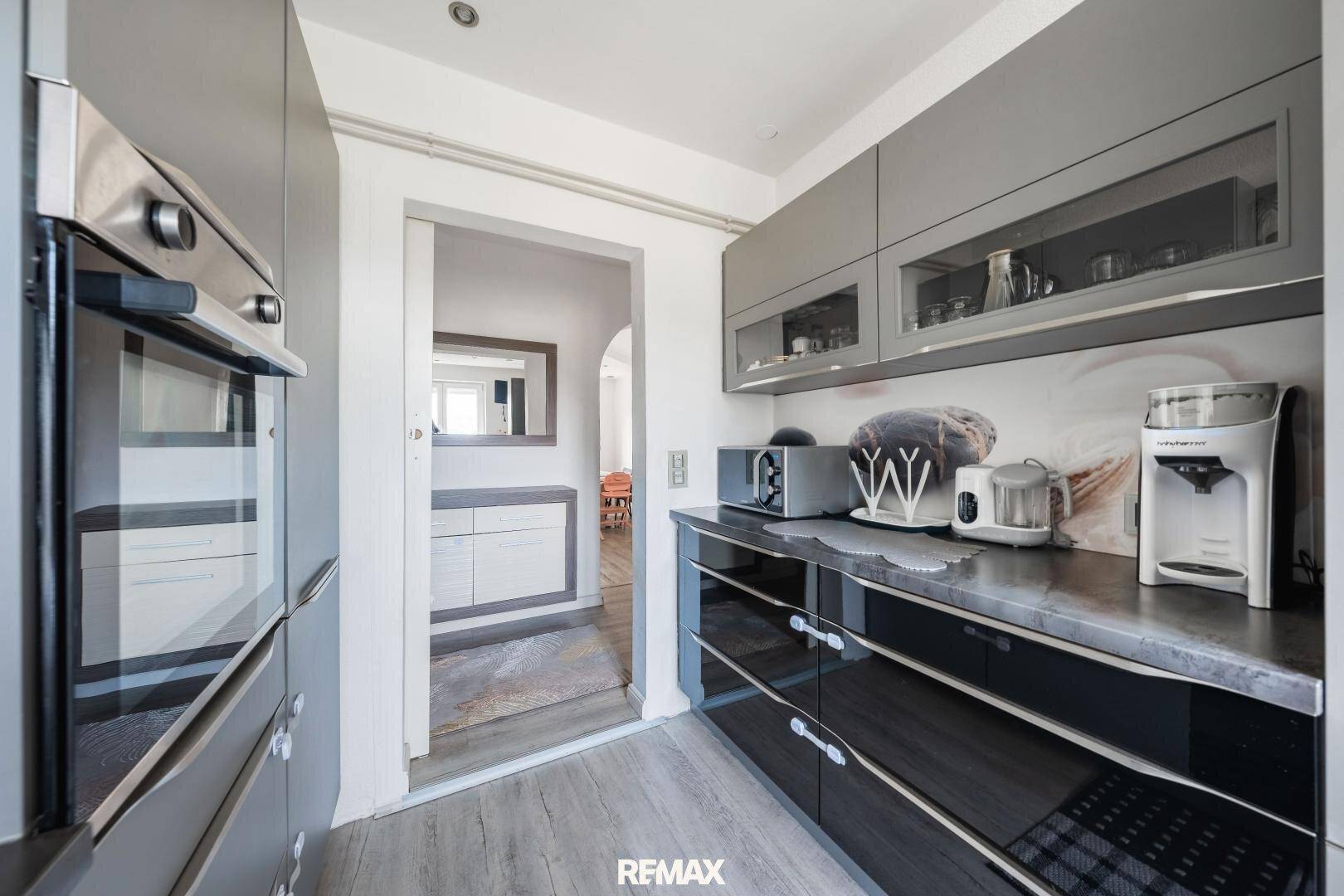 Moderne Küche mit grauen und schwarzen Hochglanzfronten, integriertem Backofen und Durchgang zum Essbereich.