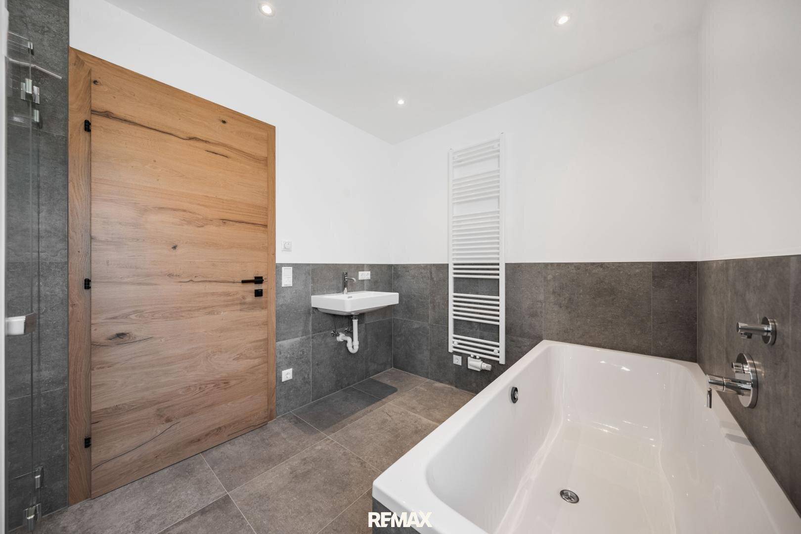 Modernes Badezimmer mit Badewanne, kleinem Waschbecken, Handtuchheizkörper und grauen Fliesen.