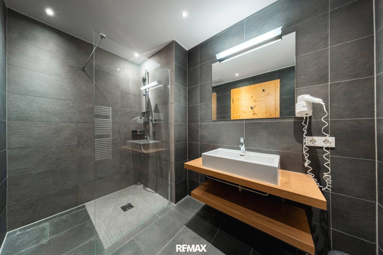 Modernes Badezimmer mit großer begehbarer Dusche, dunklen Fliesen und Holzwaschtisch.