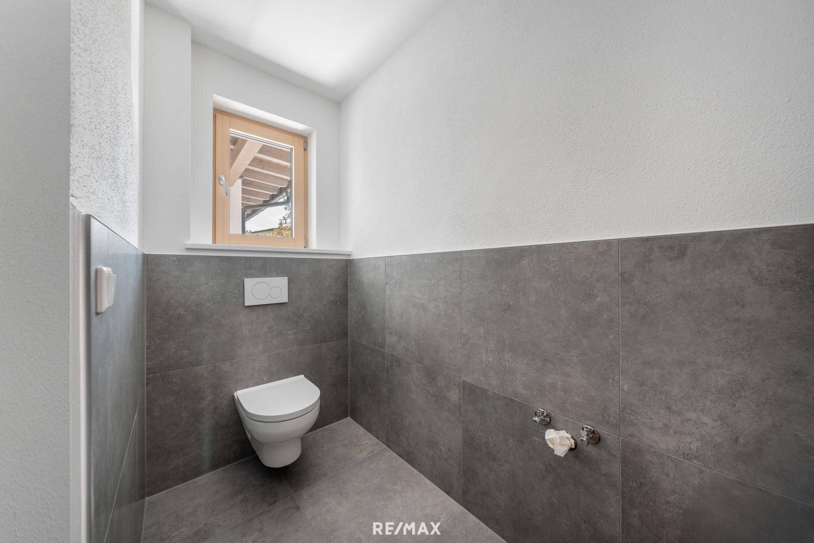 Modernes Gäste-WC mit wandhängender Toilette, grauen Fliesen und einem Fenster für Tageslicht.