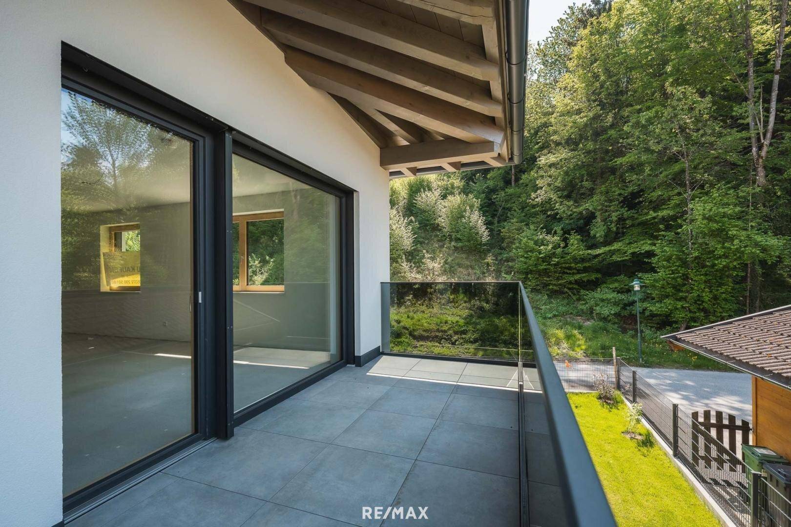Heller Balkon mit Glasgeländer, Fliesenboden und einem ruhigen Blick ins Grüne.