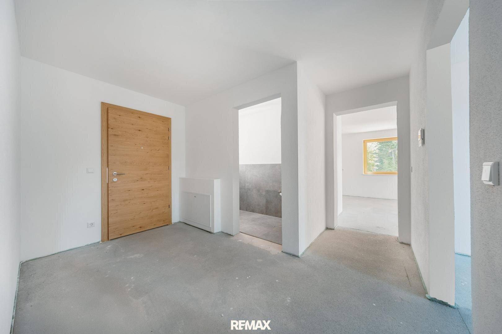 Eingangsbereich mit Holztür und unfertigem Betonboden, der zu weiteren Räumen führt.