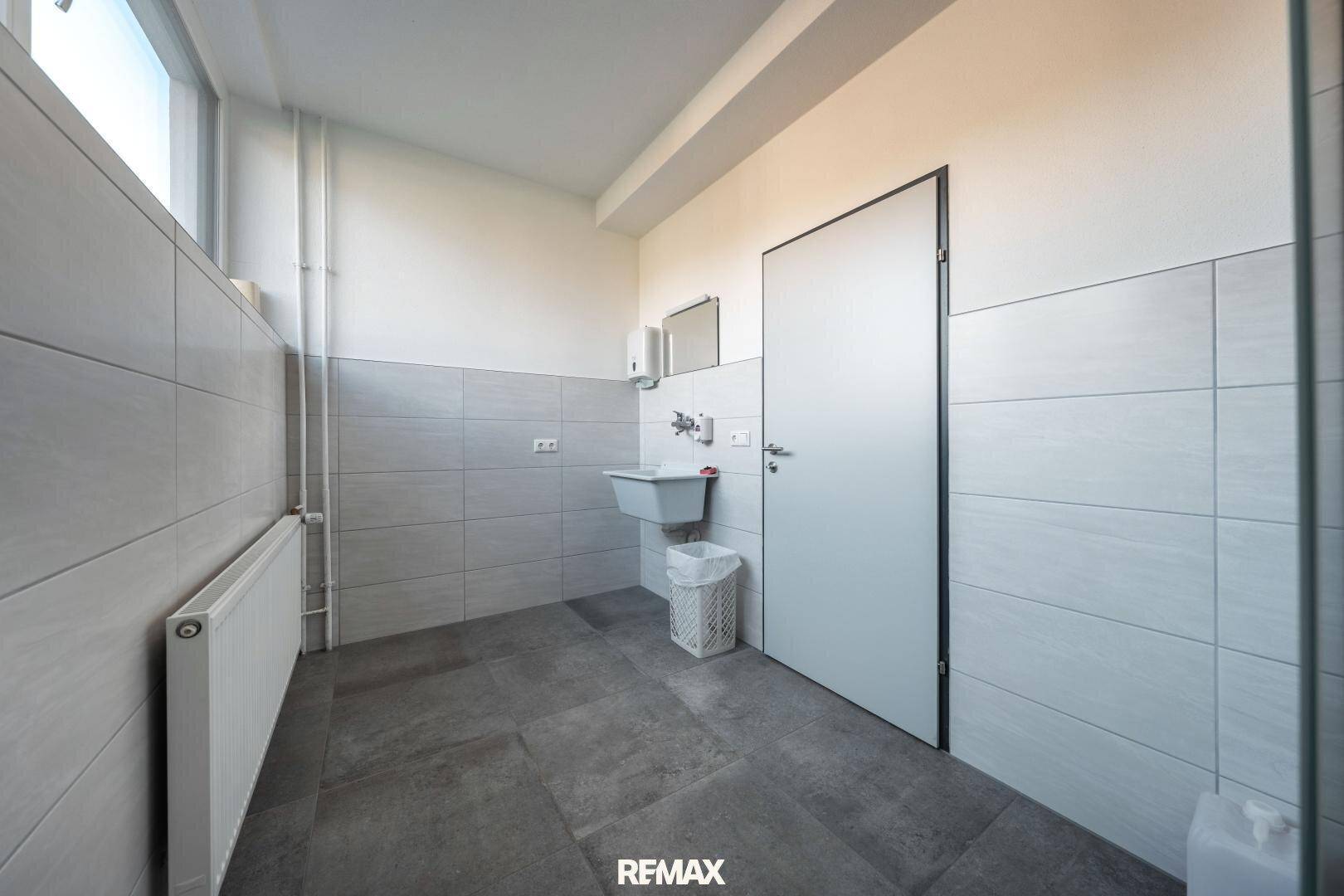 Sauberer Toilettenraum mit hellgrauen Fliesen, Waschbecken, Fenster und moderner Ausstattung.