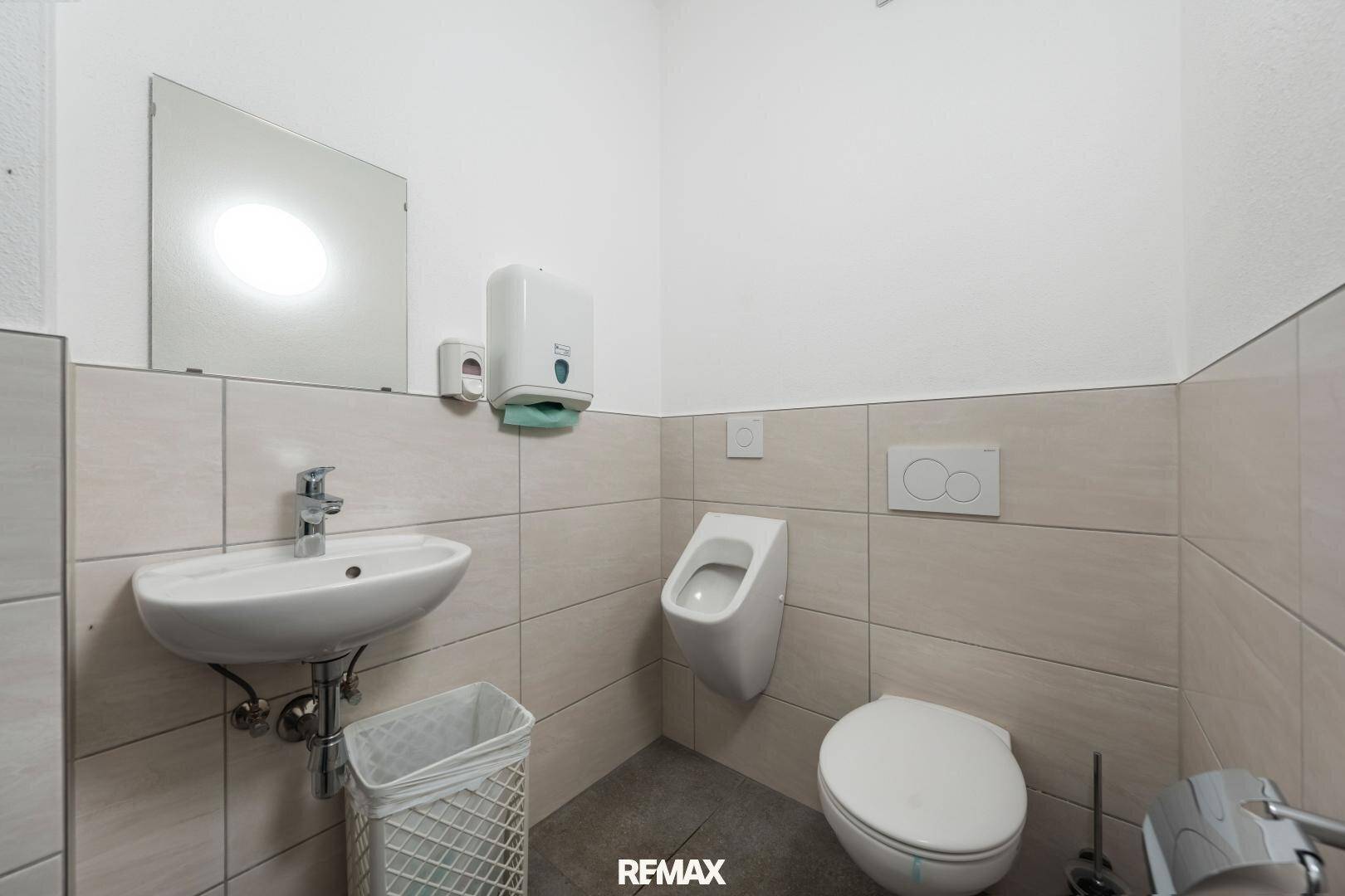 Heller Toilettenraum mit Urinal, Waschbecken und Wandfliesen in neutralen Farben.