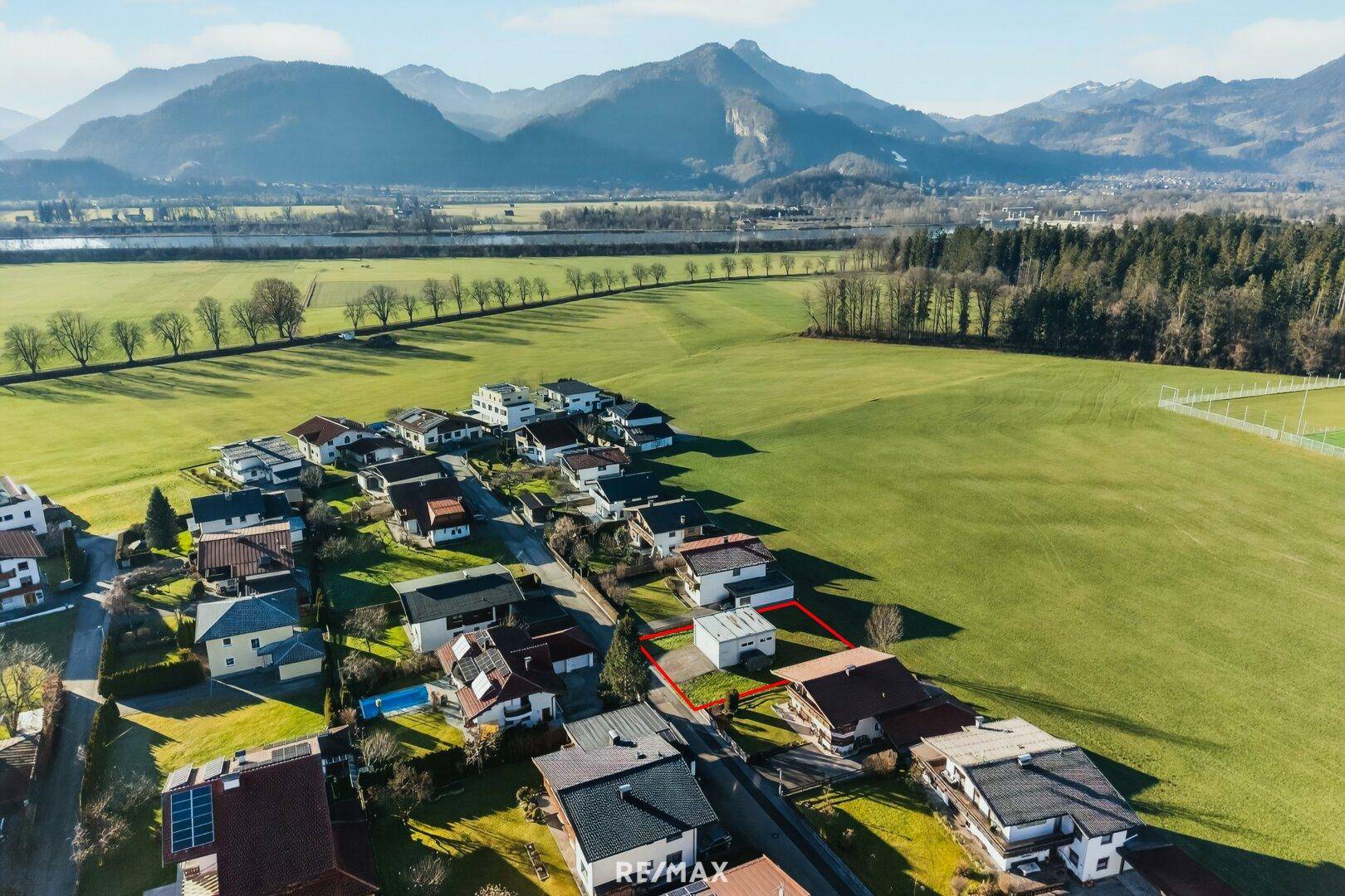 Weitläufige Luftaufnahme eines Baugrundstücks, umgeben von Feldern und Bergen, mit Sportplatz in der Nähe.