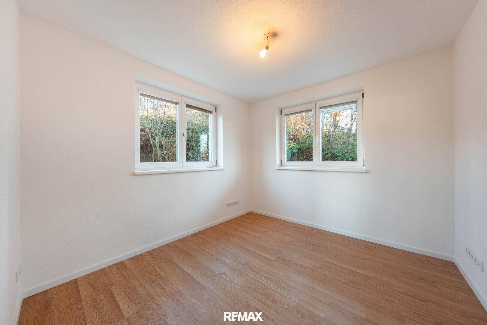 Leeres Zimmer mit Laminatboden und zwei Fenstern mit Blick ins Grüne.