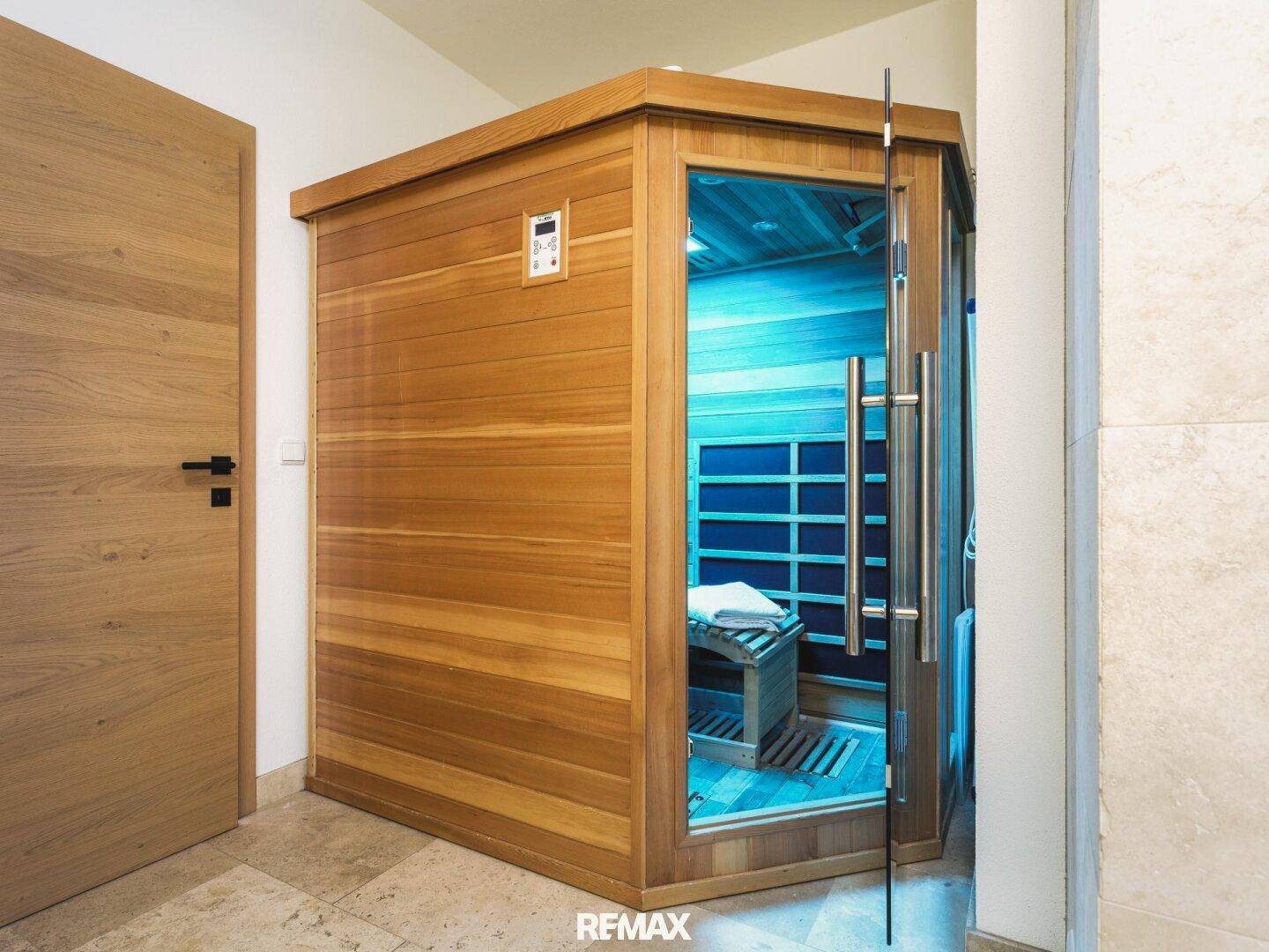Moderne Holzsauna mit Glastür und blauer Beleuchtung in einem gefliesten Bereich.