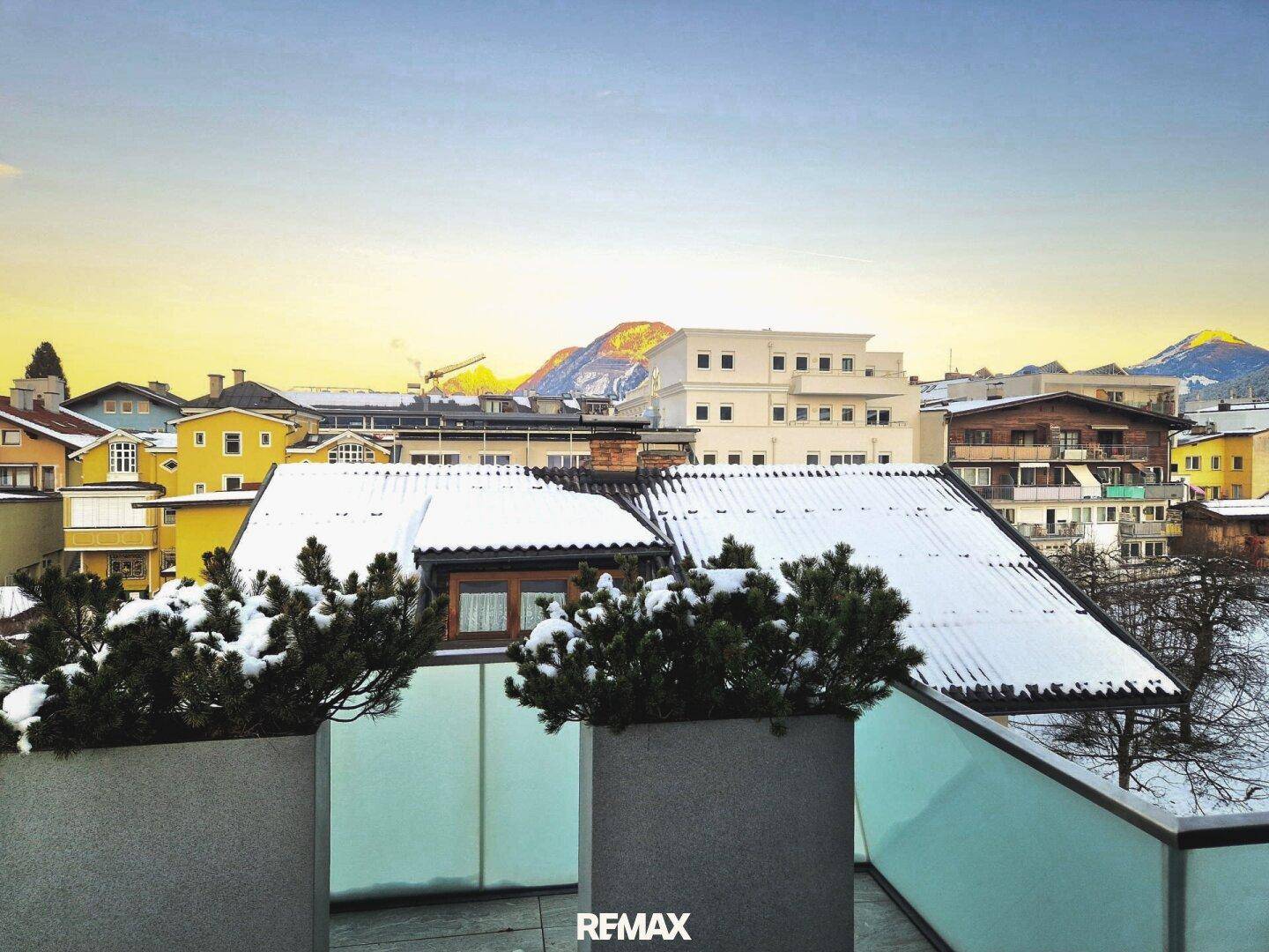 Aussicht von einer Terrasse mit schneebedeckten Dächern und Bergen im Hintergrund bei Sonnenuntergang.