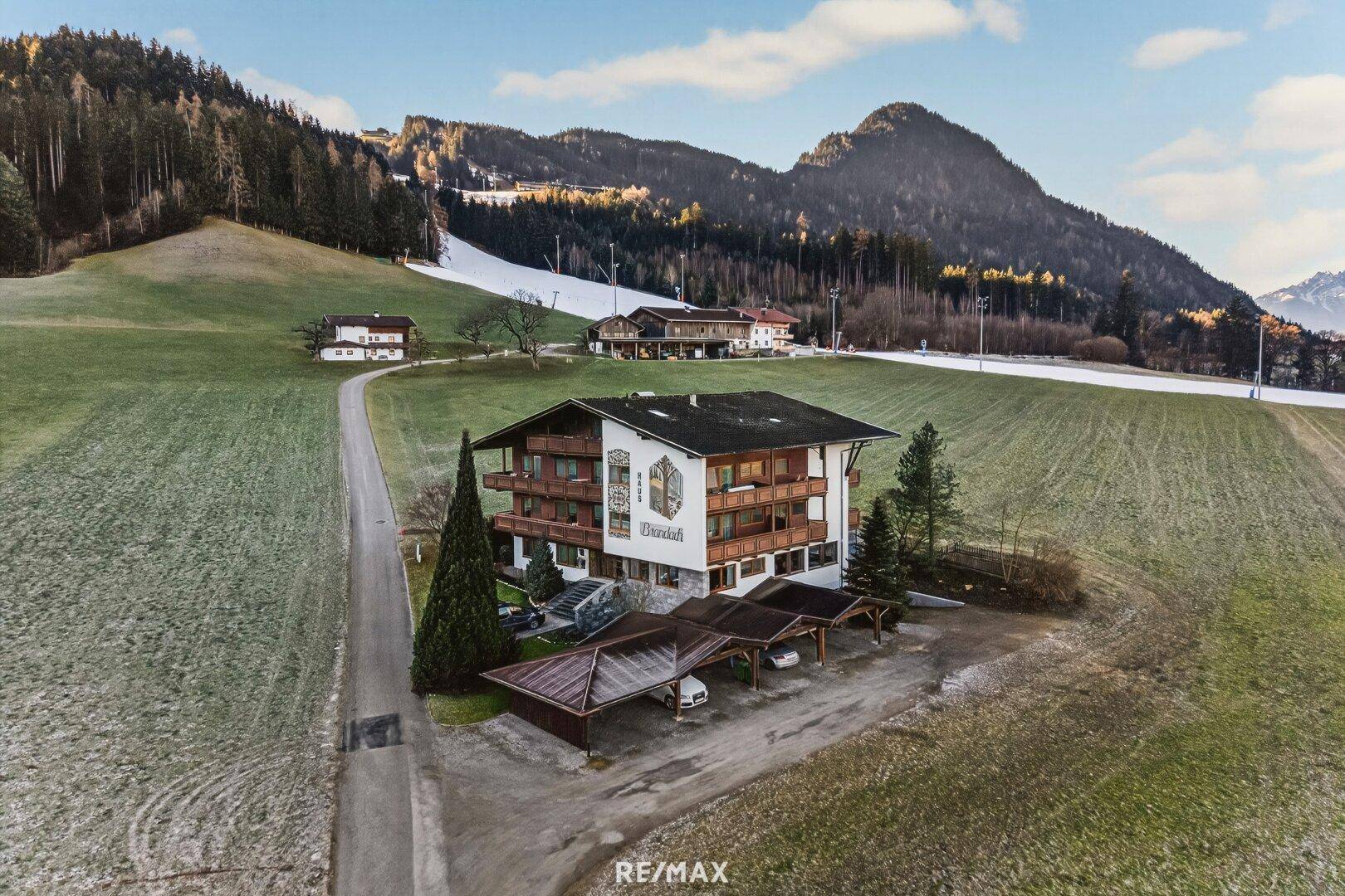Luftaufnahme des Gebäudes im alpinen Stil mit Carports und umliegenden grünen Wiesen.