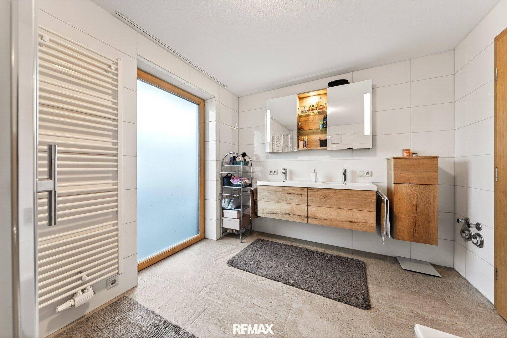 Helles Badezimmer mit Doppelwaschbecken, Spiegelschrank, beheiztem Handtuchhalter und großem Fenster.