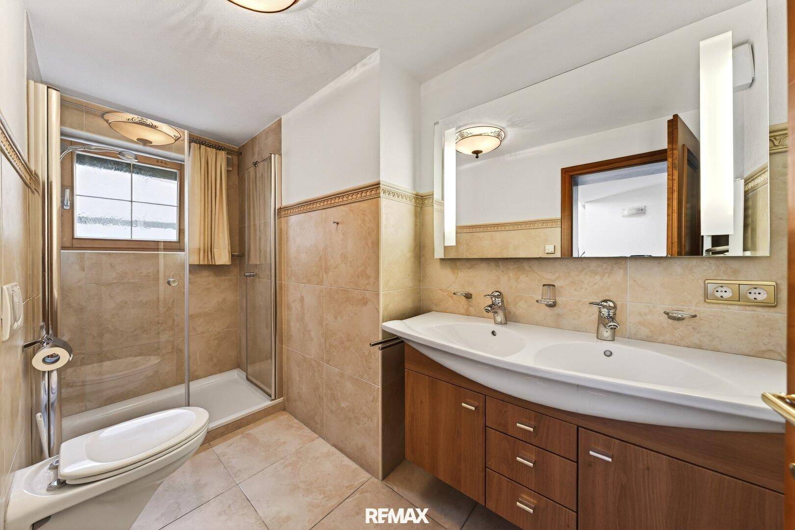 Badezimmer mit Doppelwaschbecken, Dusche und beigefarbenen Fliesen, gut beleuchtet.