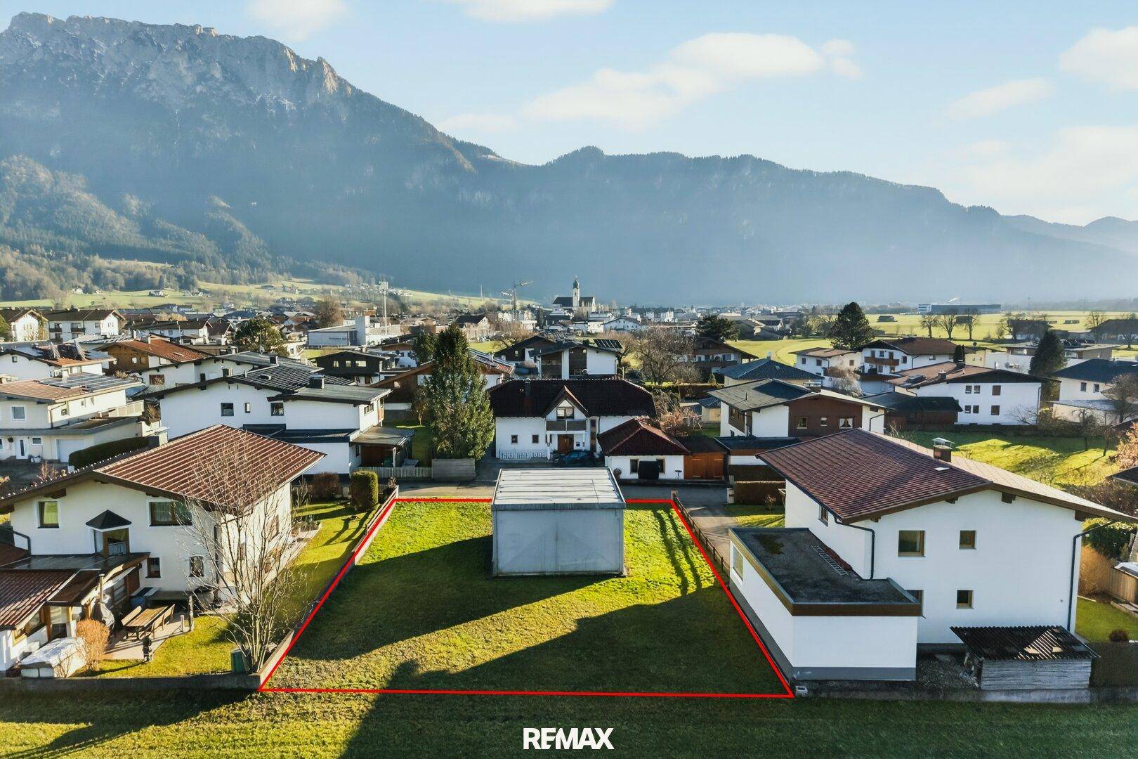 Luftaufnahme eines sonnigen Baugrundstücks in einer Siedlung mit bestehendem Nebengebäude und Bergblick.