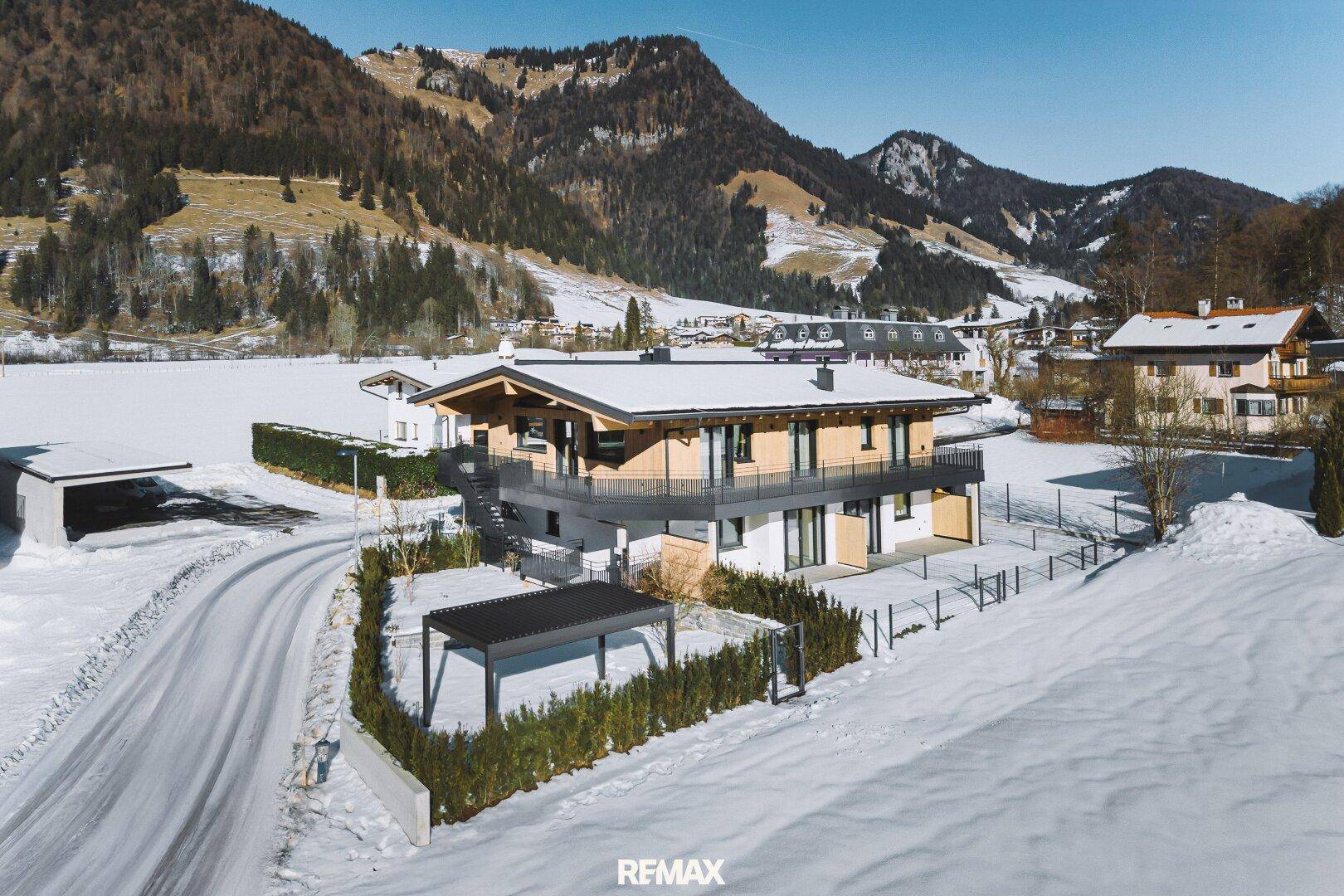 Winterliche Luftaufnahme des modernen Hauses mit verschneitem Garten, Pergola und umliegender Berglandschaft.