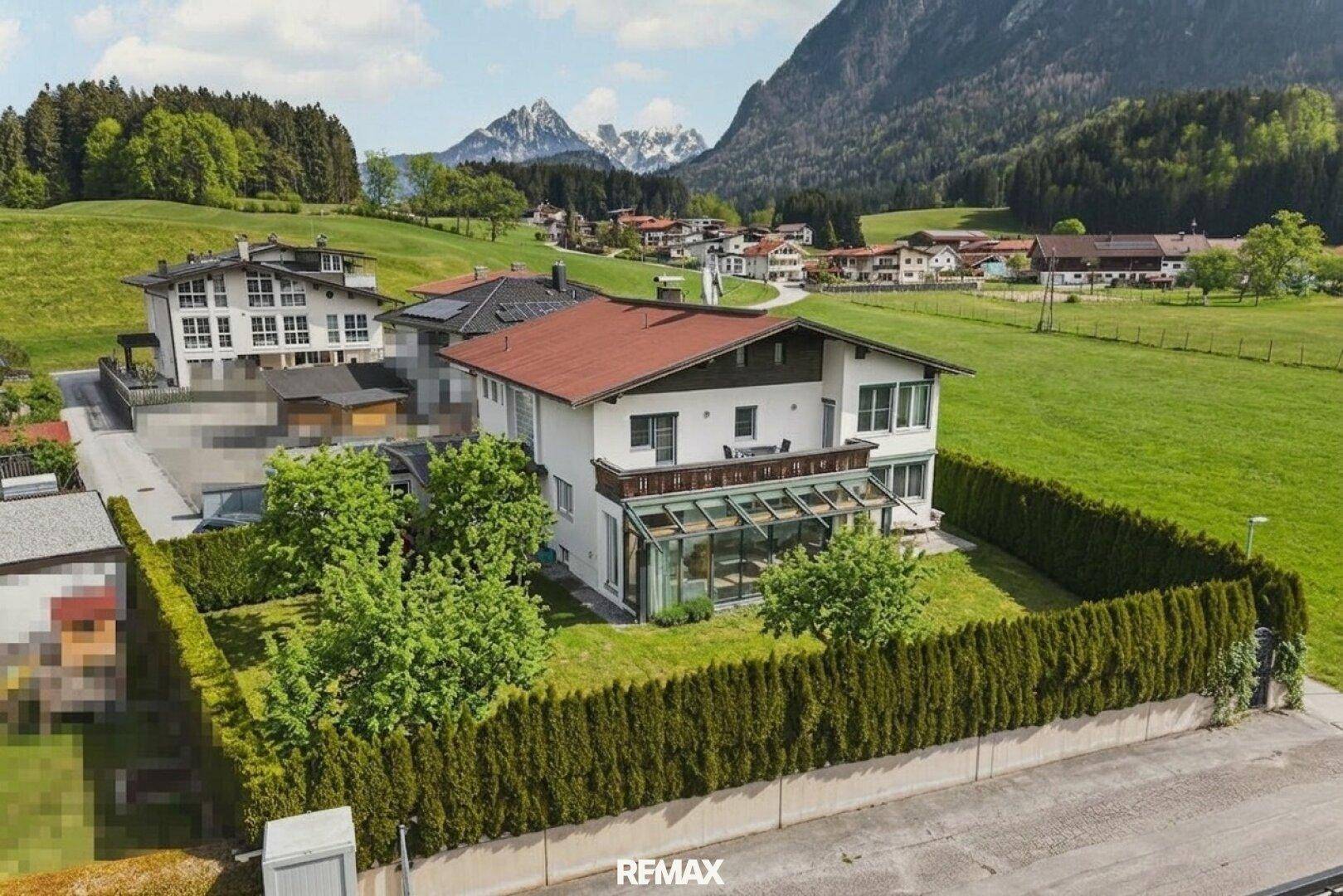 Haus mit gepflegtem Garten, hoher Hecke und atemberaubendem Blick auf die umliegenden Berge und das Dorf.
