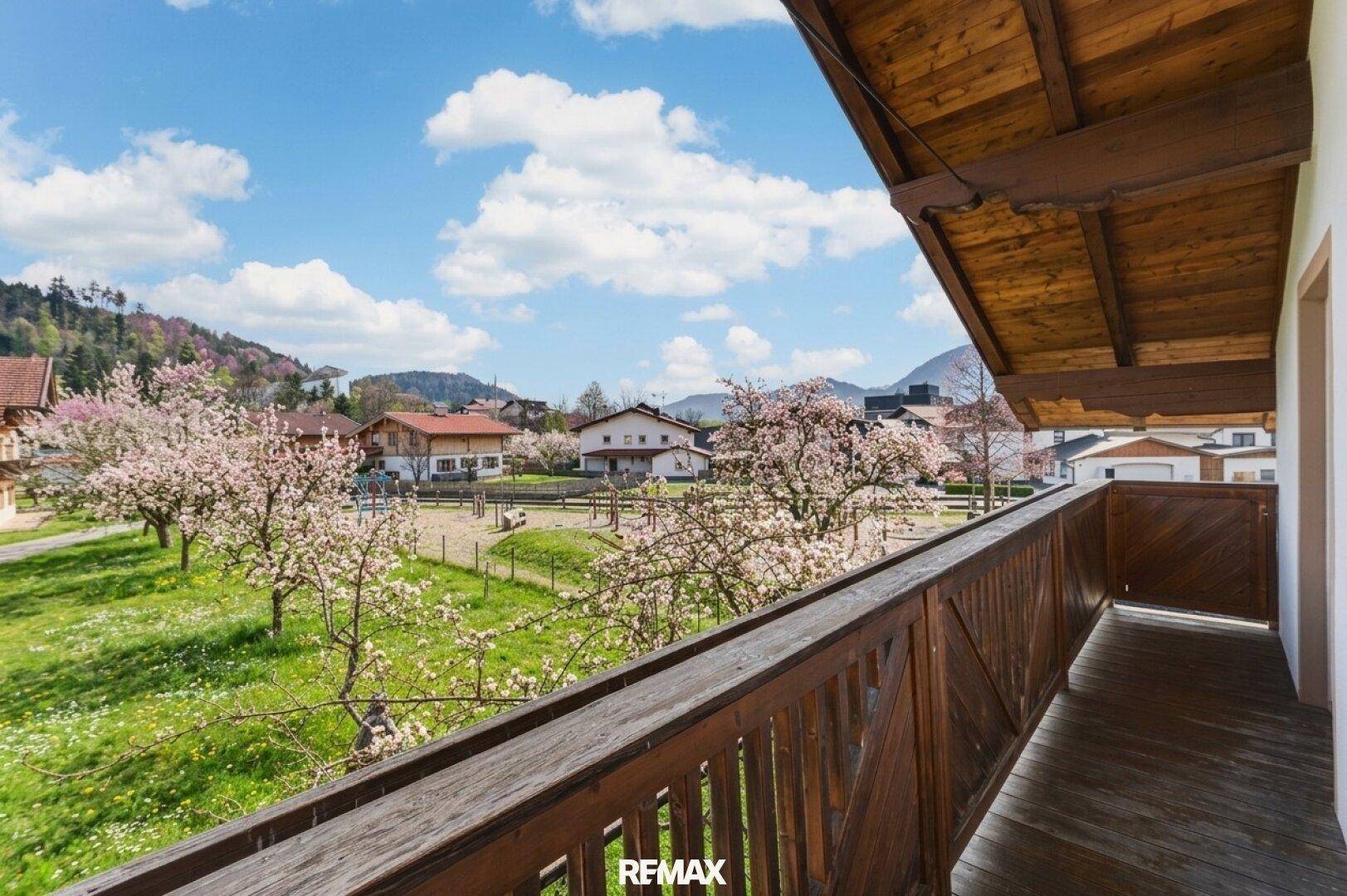 Blick vom Holzbalkon auf eine grüne Landschaft mit blühenden Bäumen und Häusern vor Bergkulisse.