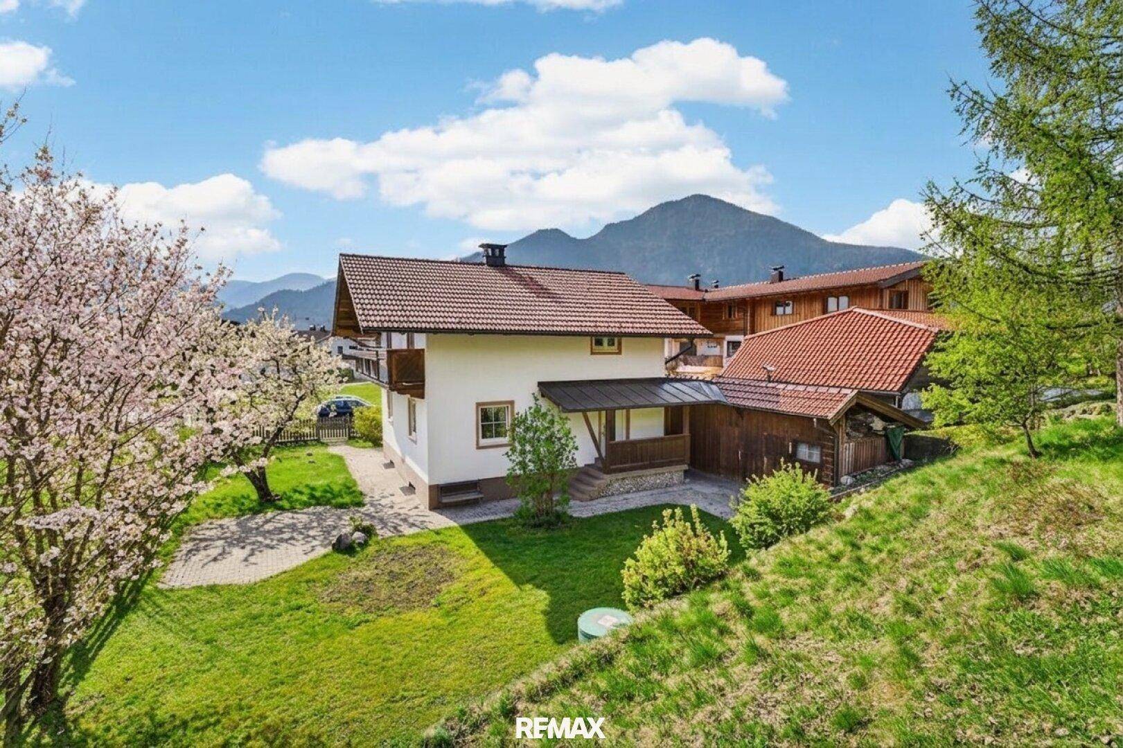 Luftaufnahme des Hauses mit rotem Ziegeldach, umgeben von grünen Wiesen und majestätischen Bergen im Hintergrund.