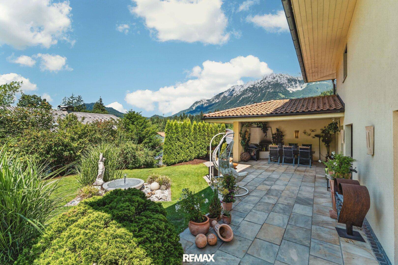 Überdachte Terrasse mit Gartenmöbeln, Brunnen und Blick auf die Berge.