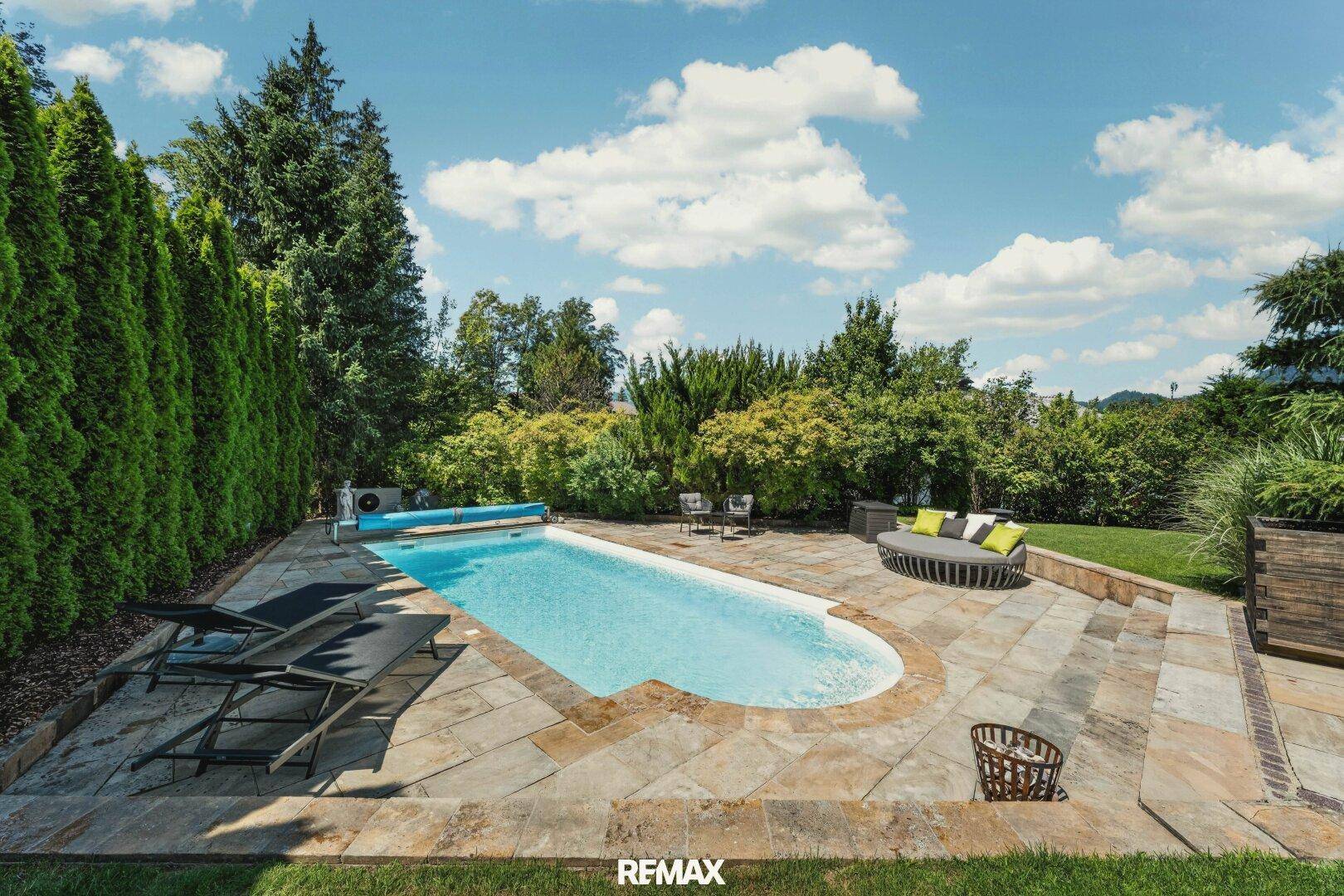 Großer Garten mit Pool, Liegeflächen und üppiger Bepflanzung unter blauem Himmel.
