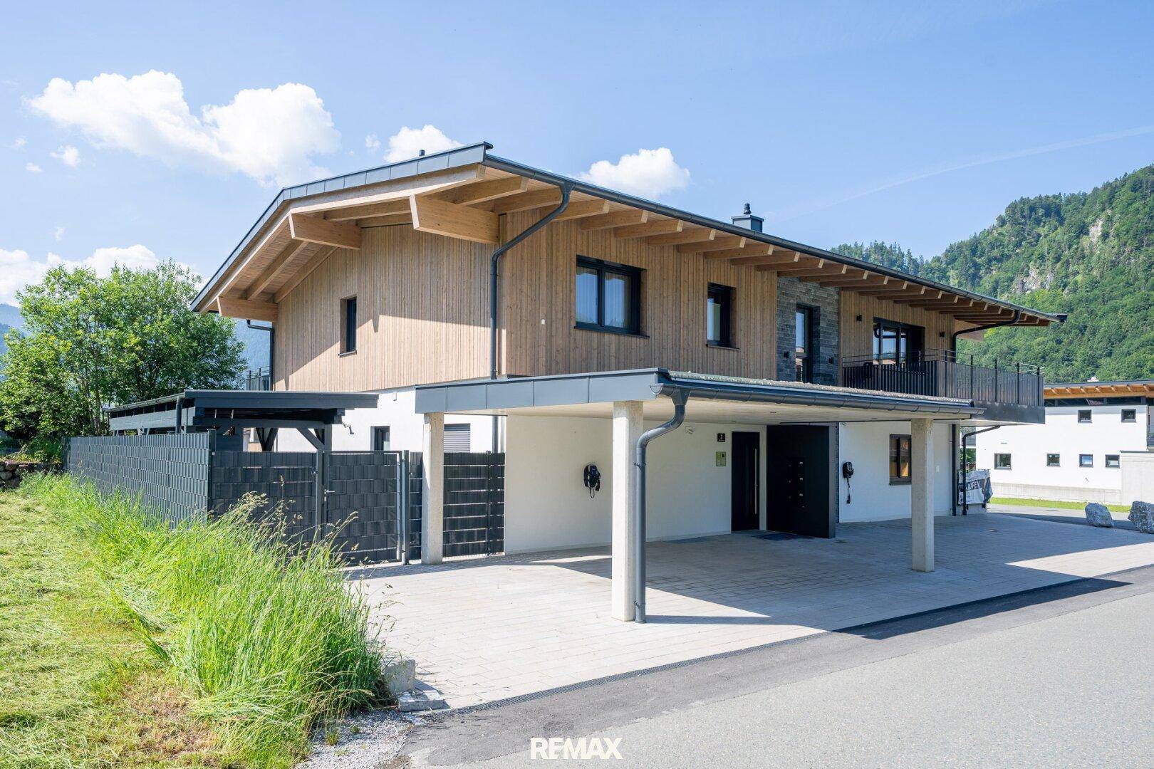 Vorderansicht des modernen Hauses mit Holzfassade, Carport und Eingangsbereich vor Bergkulisse.