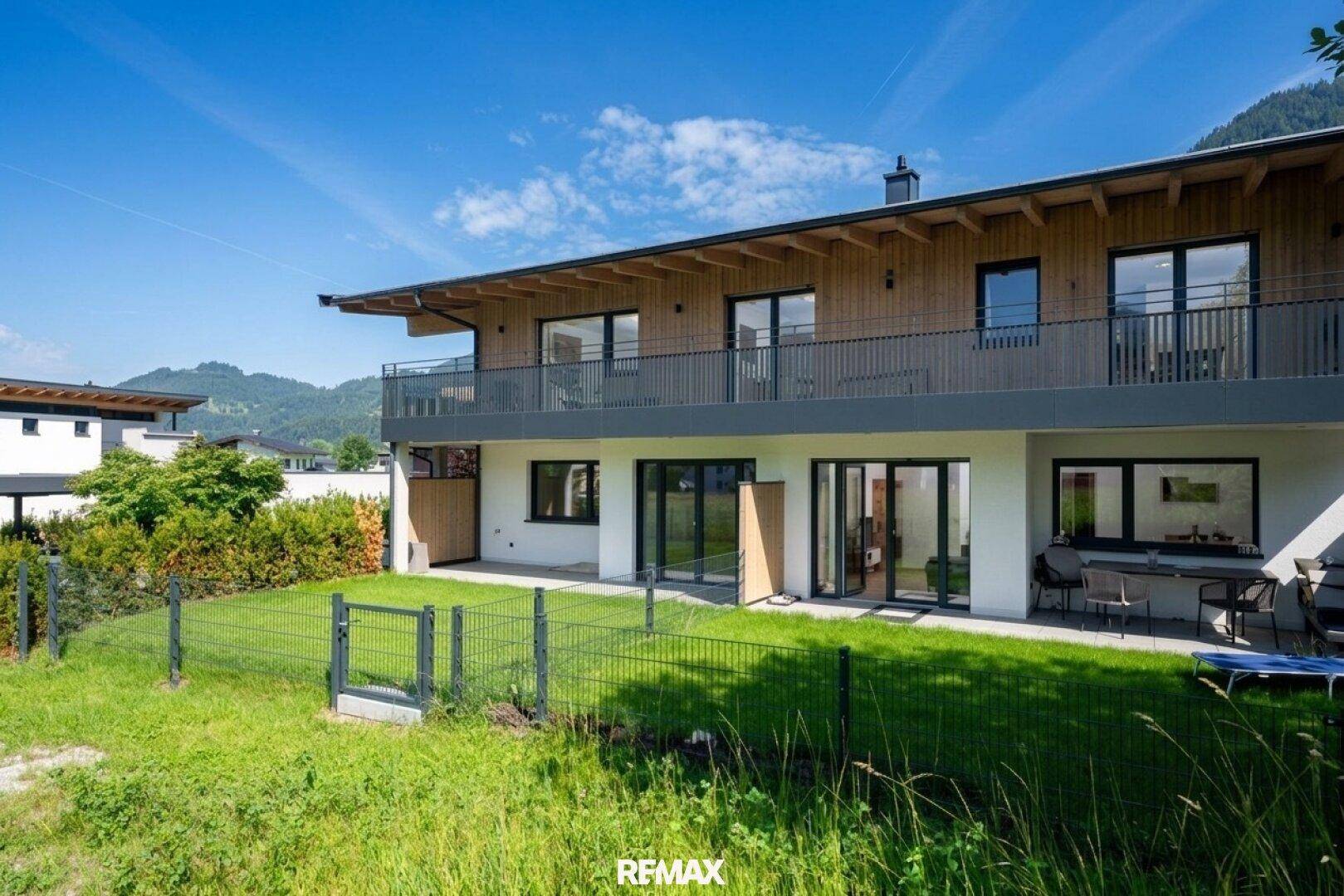 Rückseite des modernen Hauses mit Terrasse, Rasenfläche und Balkon im Obergeschoss, umgeben von Natur.
