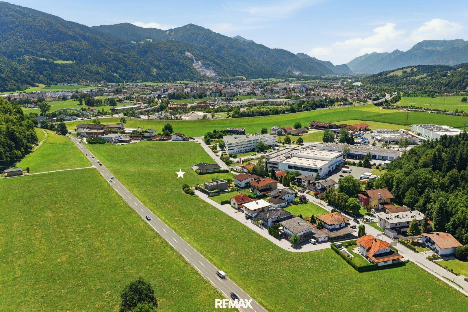 Luftaufnahme einer ländlichen Umgebung mit Wohngebäuden, Feldern und einer Straße, umgeben von Bergen.