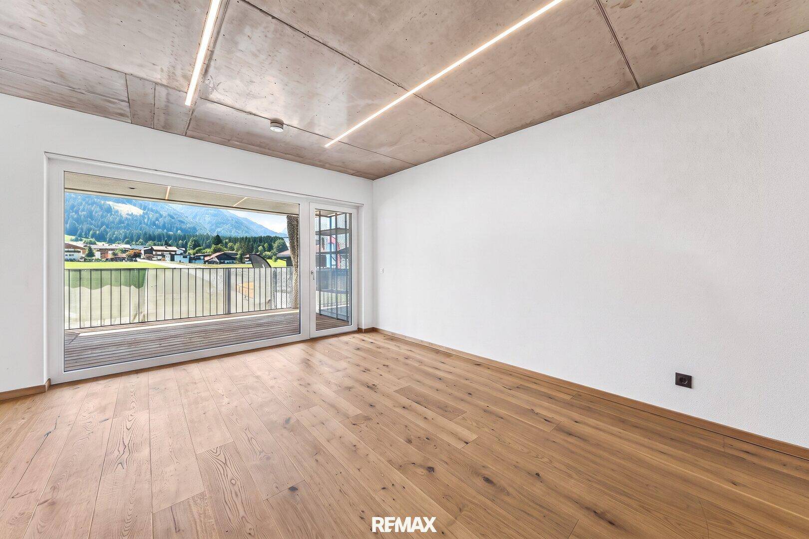 Heller Raum mit Holzboden, Sichtbetondecke und Zugang zu einem Balkon mit Bergblick.
