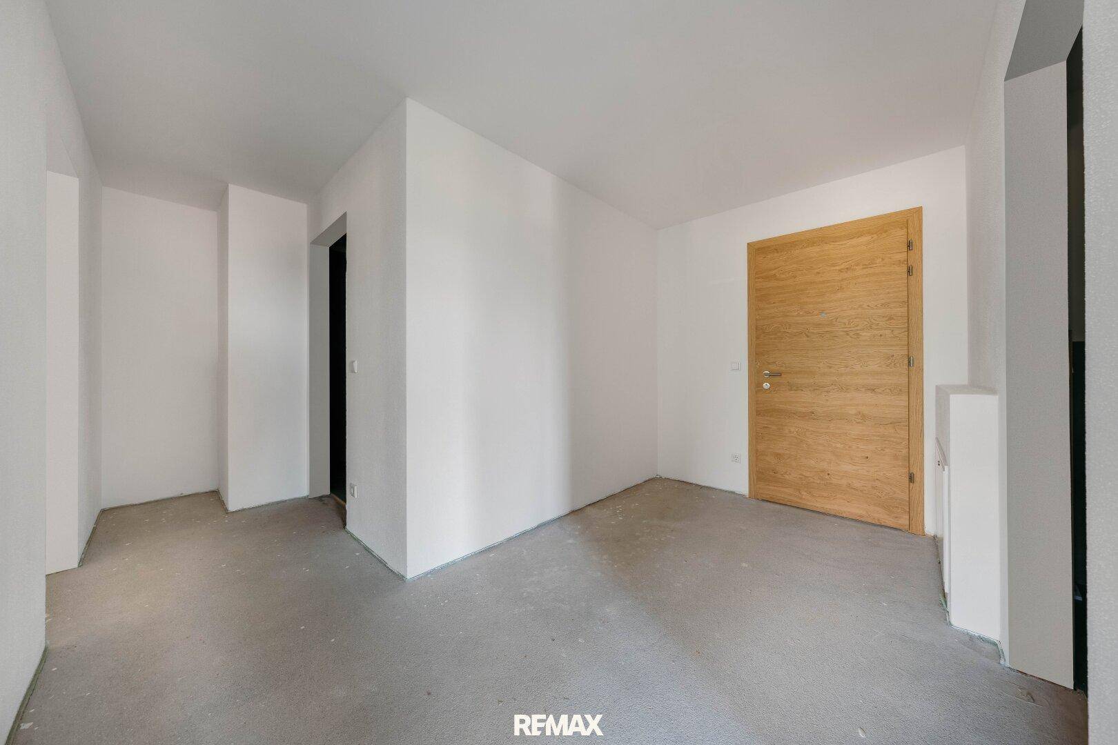Unfertiger Innenraum mit weißen Wänden, Betonboden und einer Holztür, bereit für den Ausbau.