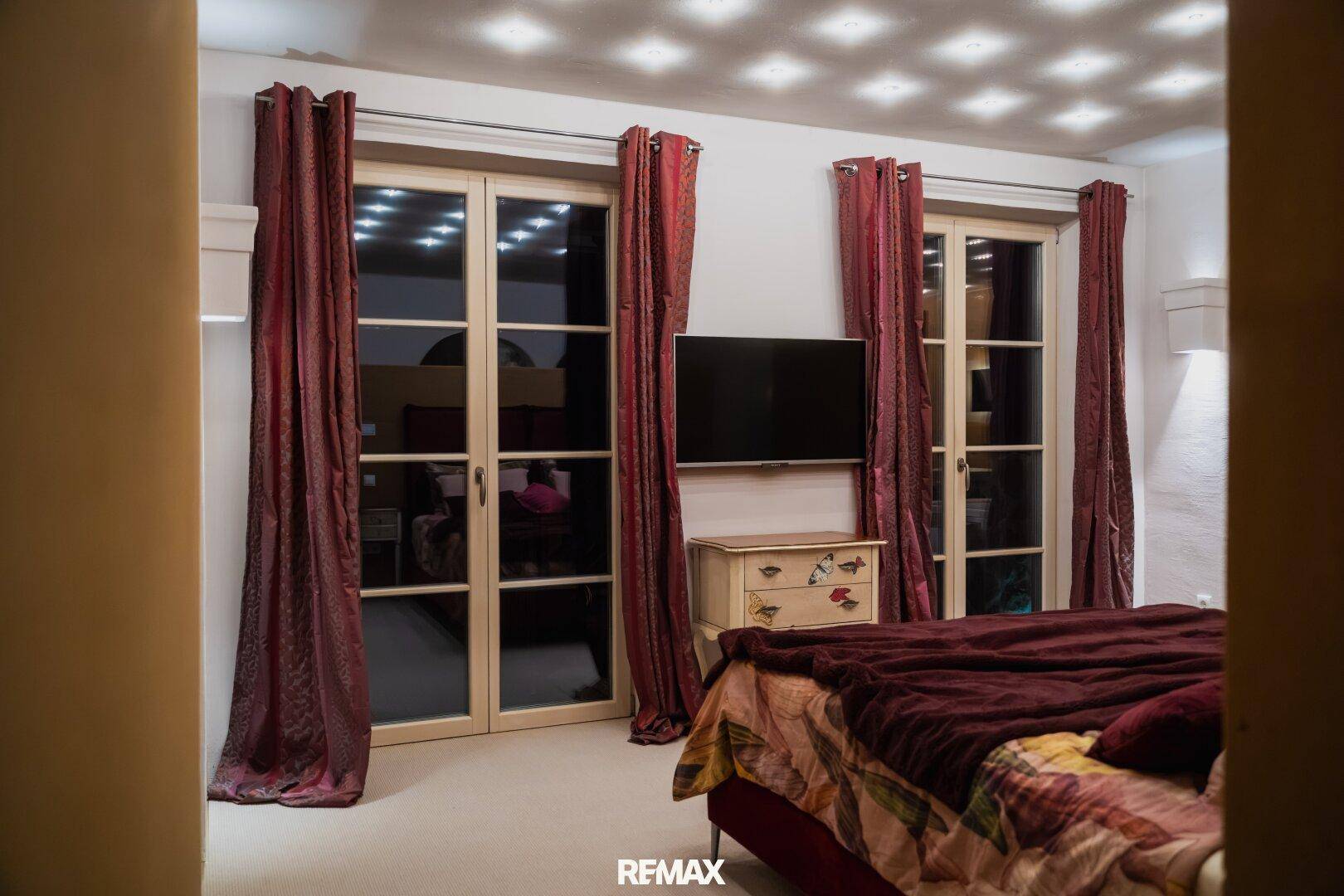Schlafzimmer mit großen Fenstertüren, rotem Bett und passenden Vorhängen, die Gemütlichkeit schaffen.