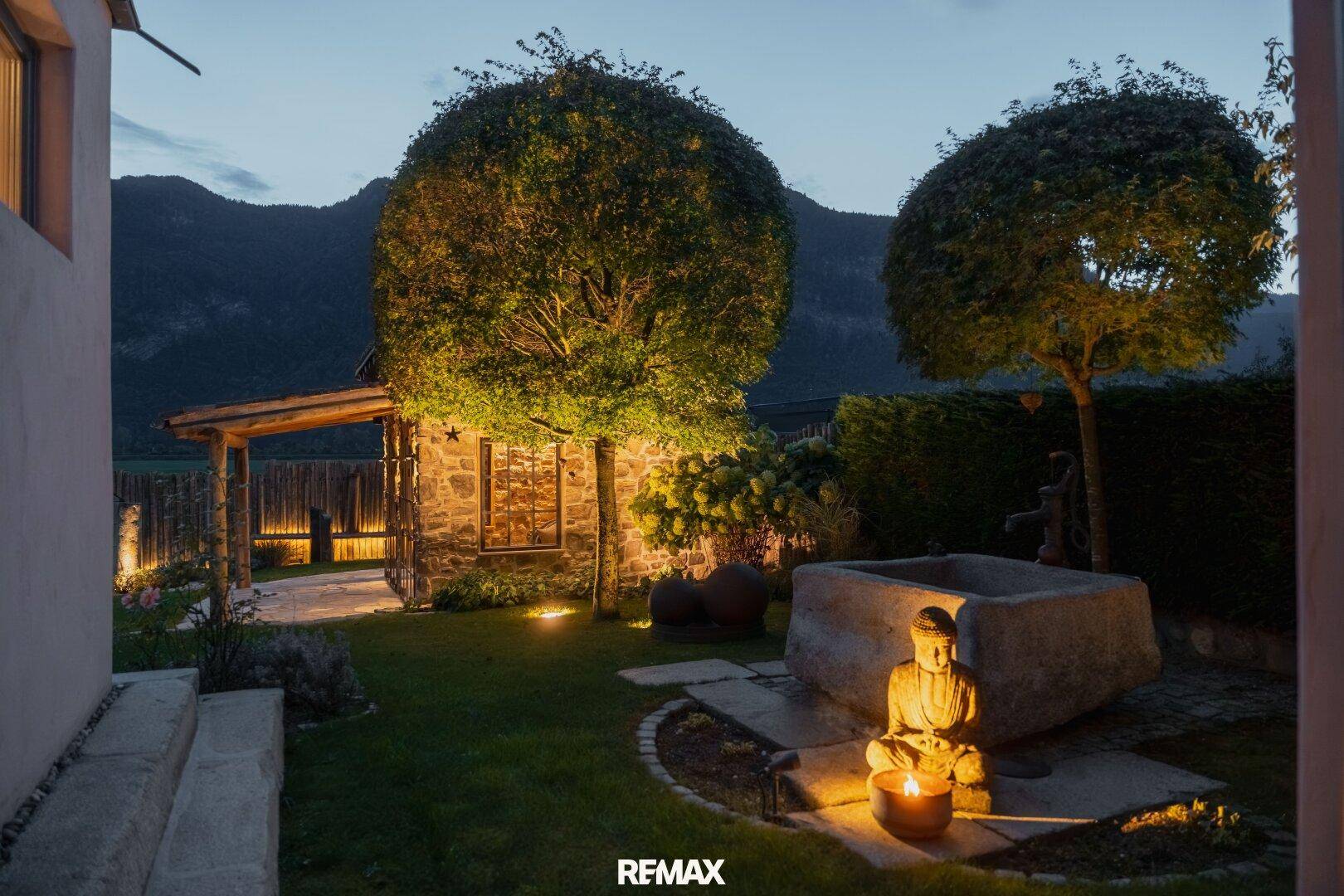Idyllischer Gartenbereich mit Buddha-Statue, Wasserbecken und gepflegten Bäumen bei Abendbeleuchtung.