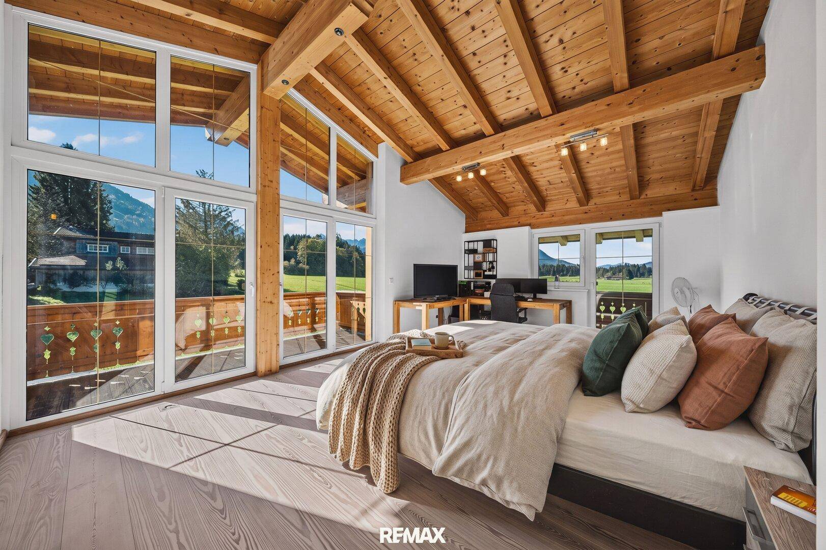Geräumiges Schlafzimmer mit Holzbalkendecke, großen Fenstern und Zugang zum Balkon.