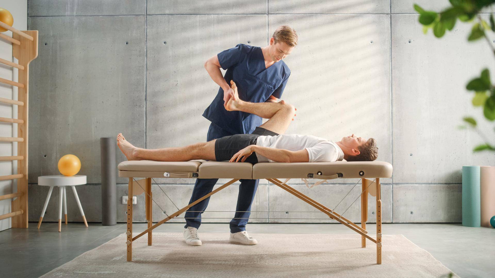 Heller Therapieraum mit minimalistischer Einrichtung, Sprossenwand und Betonwänden für physiotherapeutische Behandlungen.