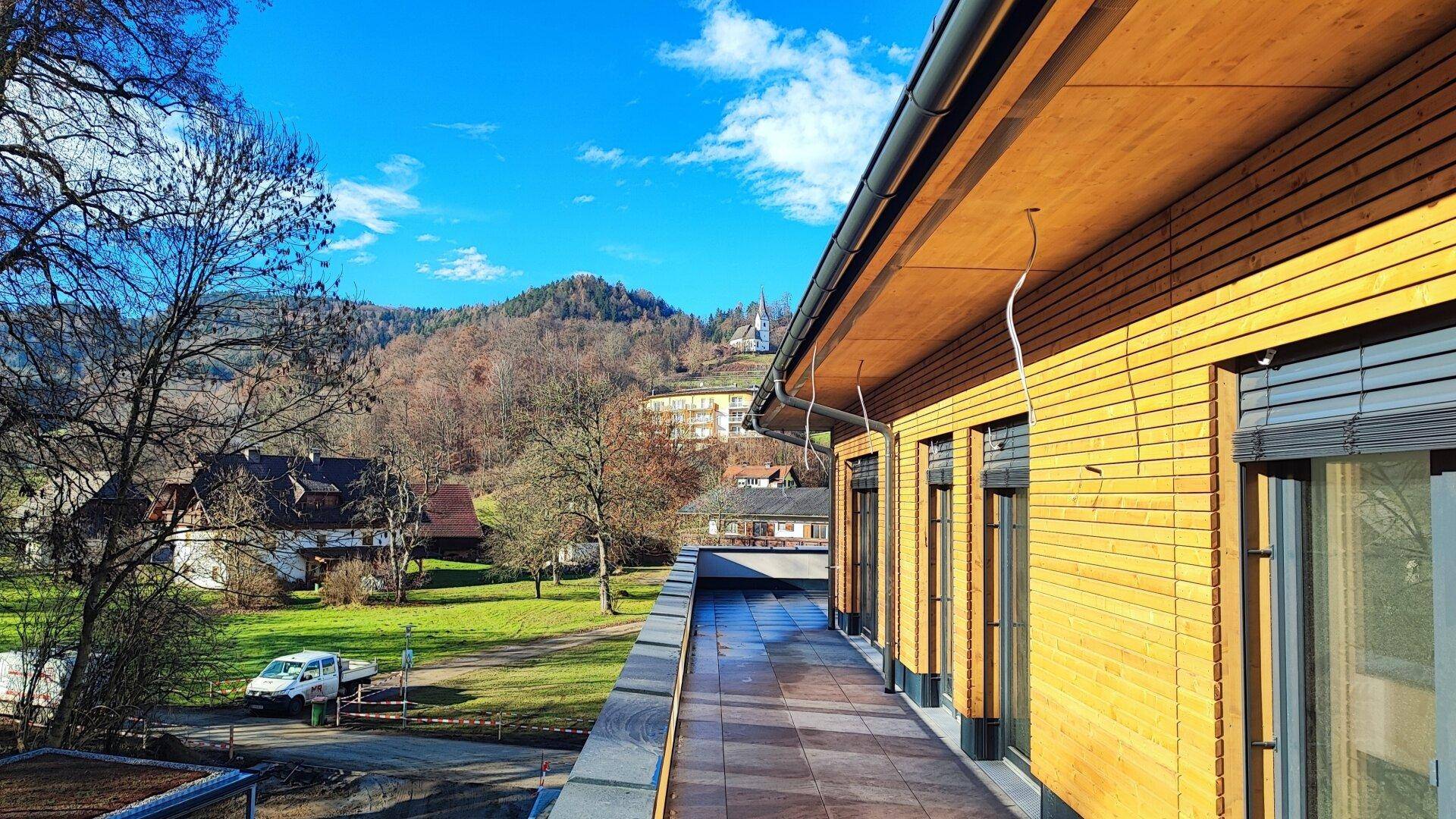 Lange Terrasse mit Fliesenboden und Ausblick auf die grüne Umgebung und entfernte Gebäude.
