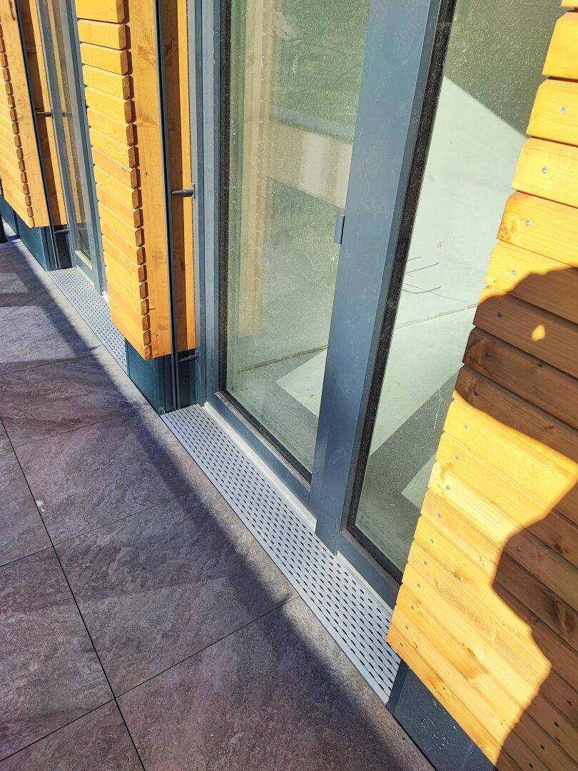Nahaufnahme des Fliesenbodens der Terrasse mit Entwässerungsrinne und großen Fenstern.