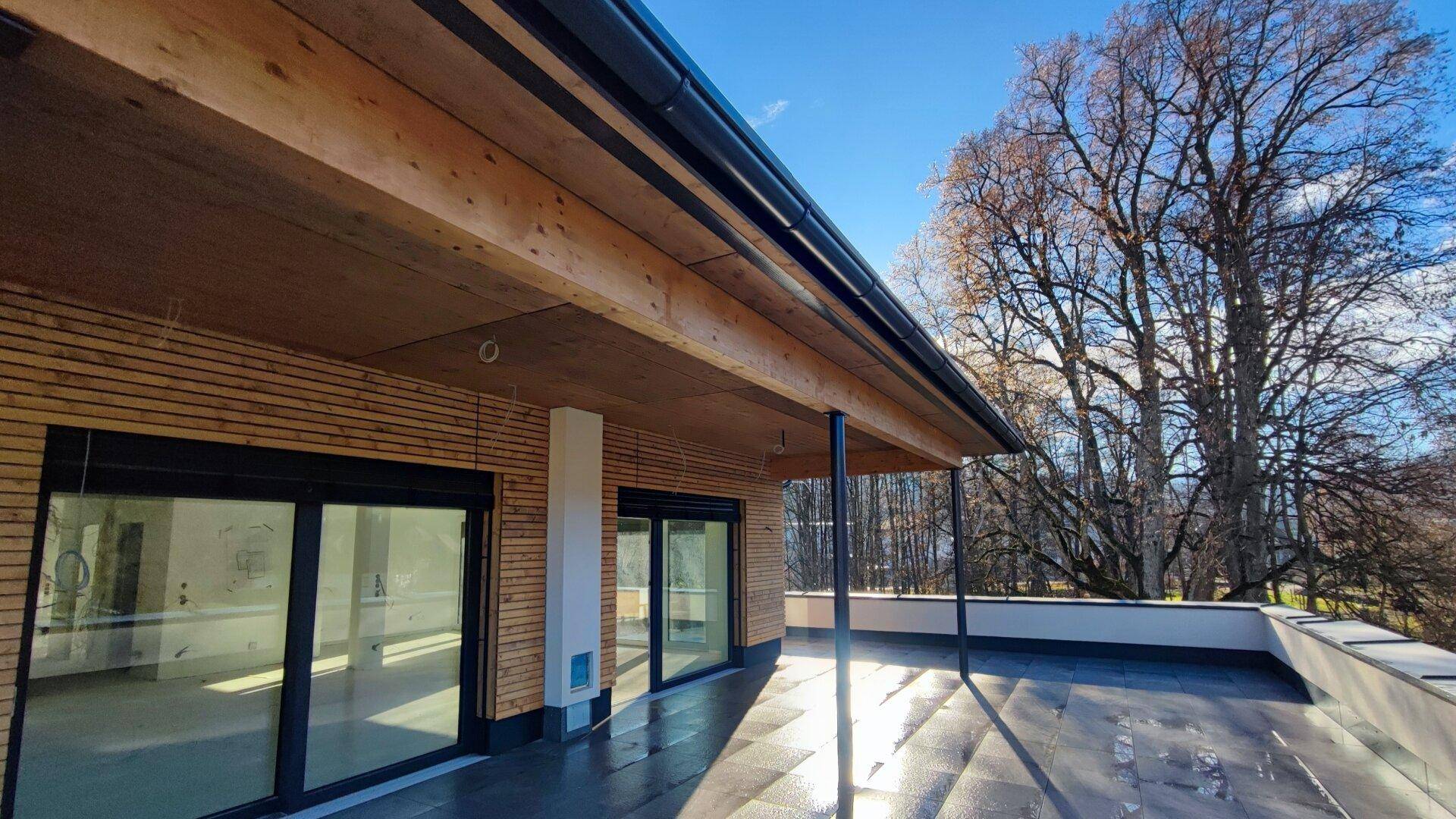 Überdachte Terrasse mit Fliesenboden und Zugang zu den Innenräumen, umgeben von Bäumen.