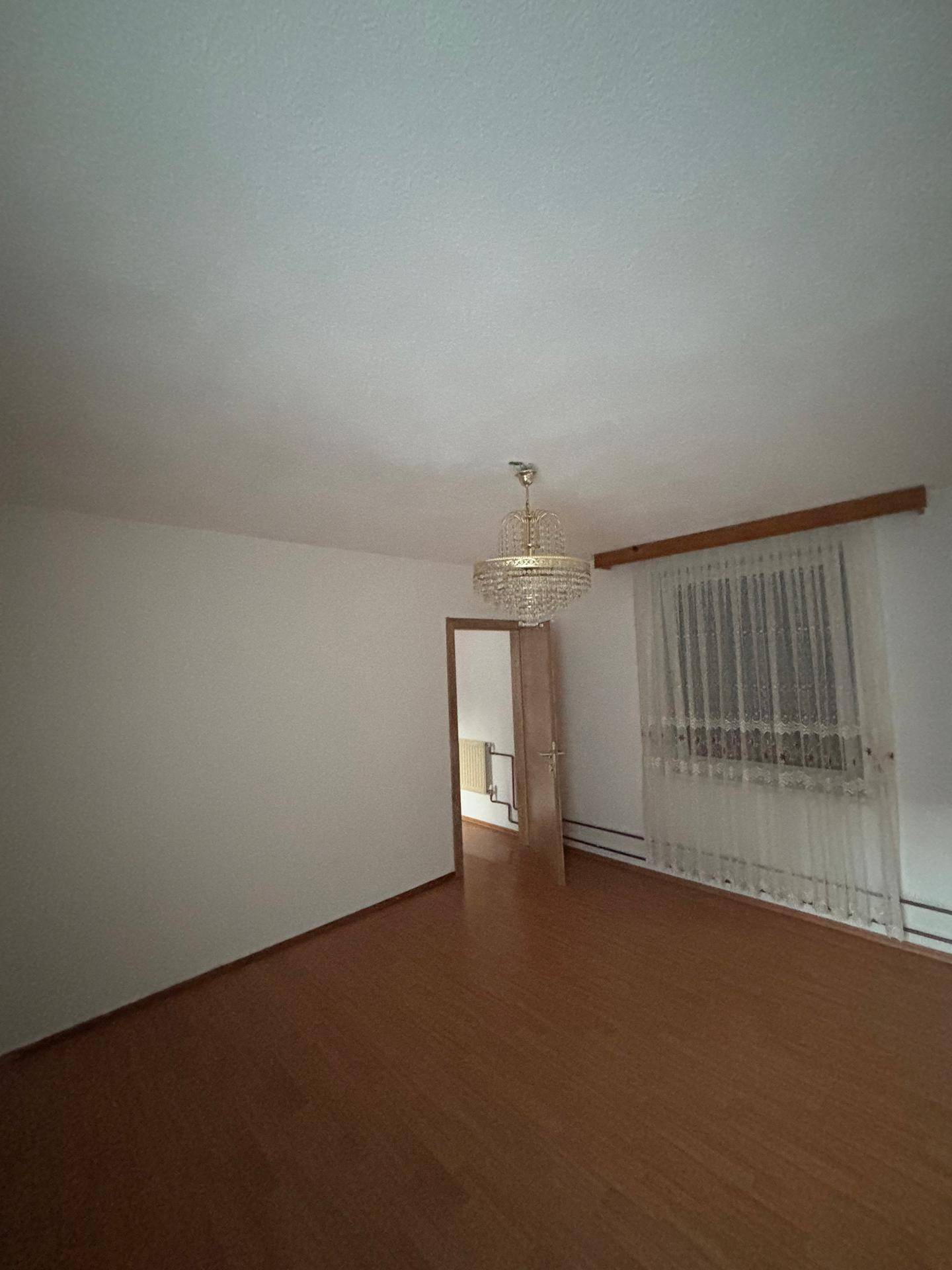 Leeres Zimmer mit Laminatboden, Fenster mit Vorhängen und einem Kronleuchter.