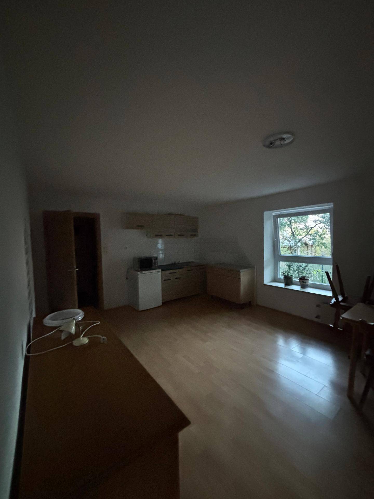 Dunkler Wohnraum mit Holzboden, Fenster und einer kleinen Küchenzeile in der Ecke.