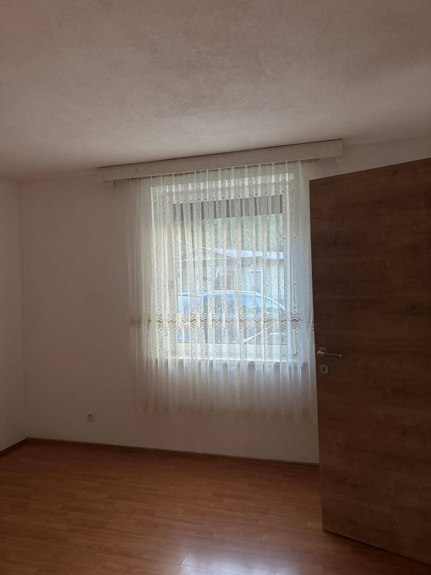 Helles Zimmer mit Laminatboden und einem Fenster mit transparenten Vorhängen.