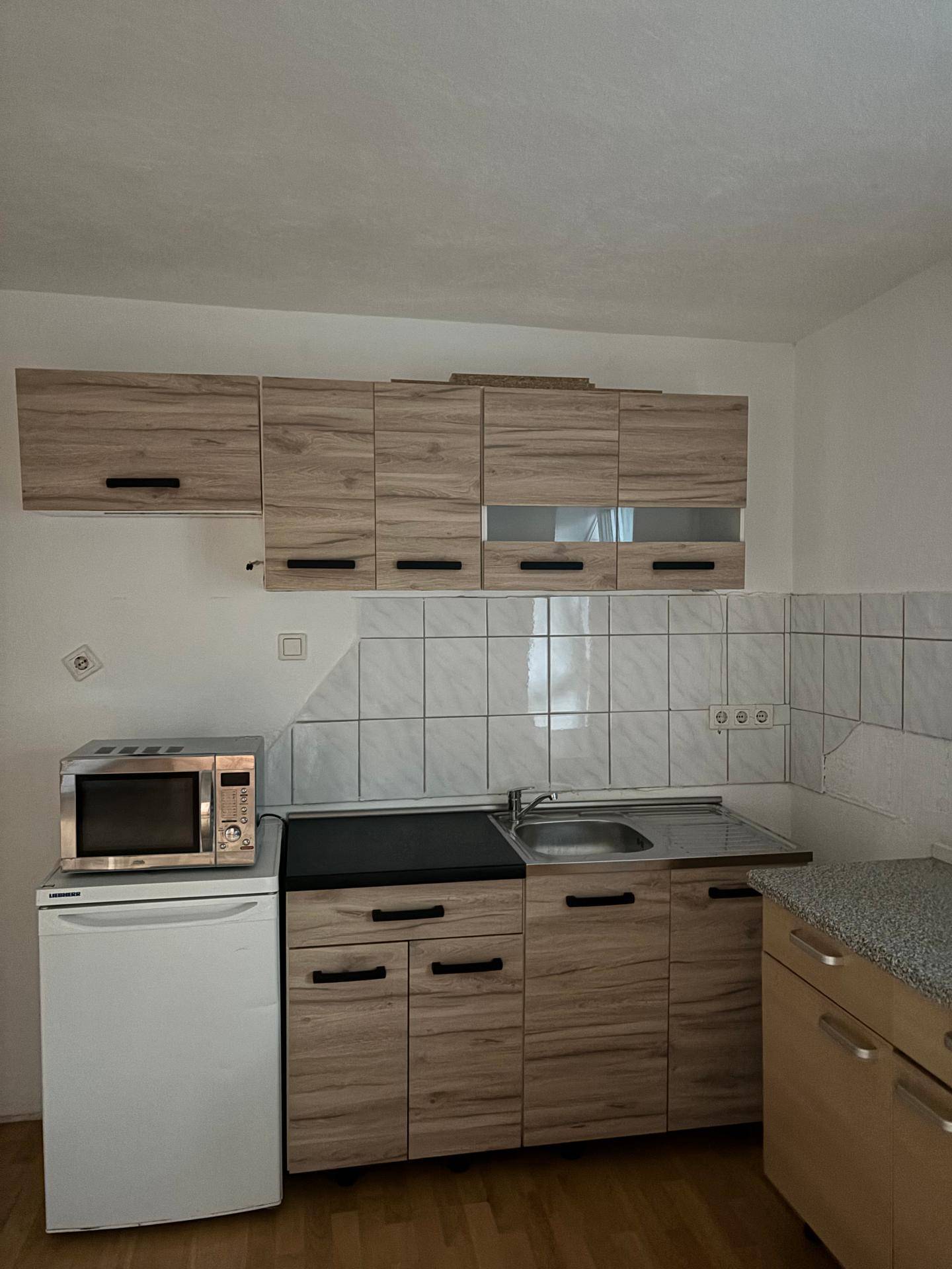 Kleine Küchenzeile mit Holzschränken, Spüle, Mikrowelle und Kühlschrank.