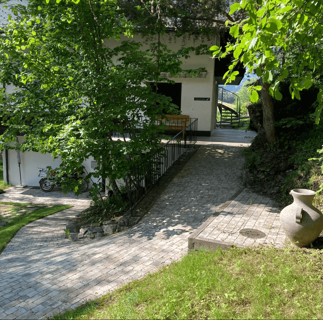 Gepflasterter Weg führt zu einem Haus, umgeben von üppigem Grün und Bäumen.
