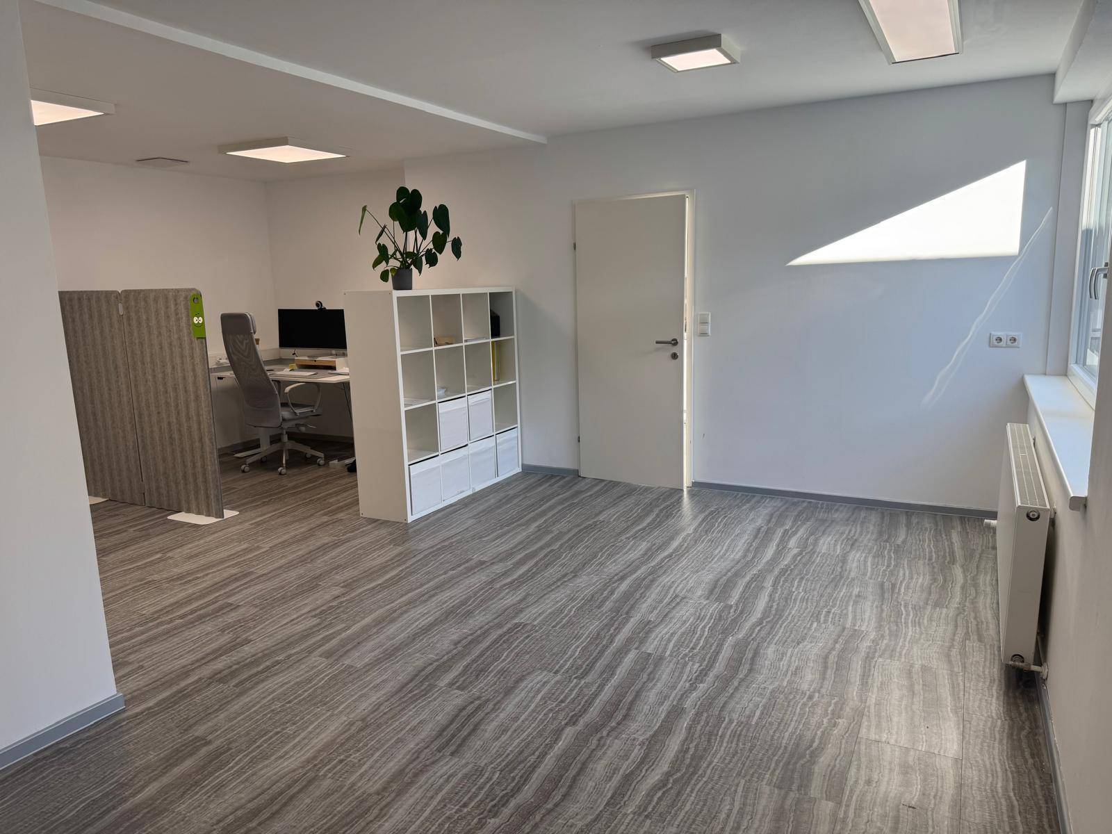 Offener Bürobereich mit Vinylboden, weißen Wänden und einem Arbeitsplatz im Hintergrund.