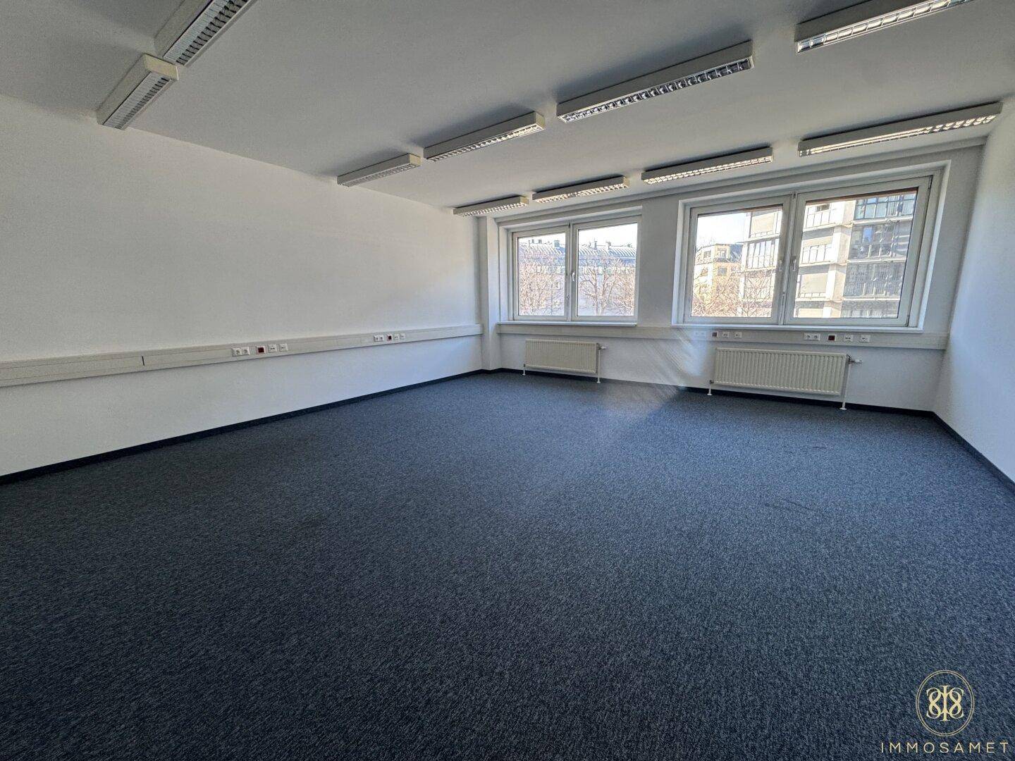Geräumiges, leeres Büro mit Teppichboden und einer breiten Fensterfront, die viel Tageslicht bietet.