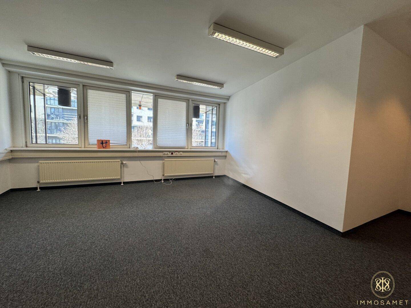 Leeres Büro mit Teppichboden, großen Fenstern und Heizkörpern unter den Fensterbänken.