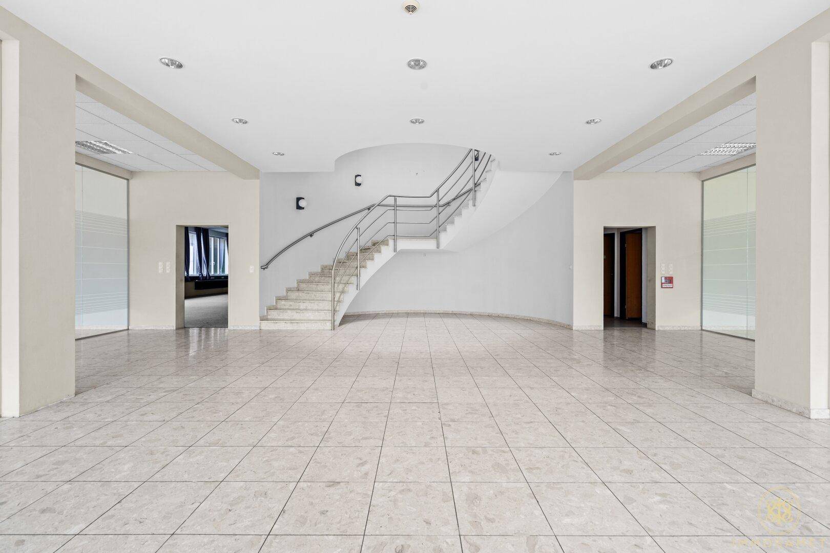 Großzügige Eingangshalle mit geschwungener Treppe, Fliesenboden und hohen Decken.