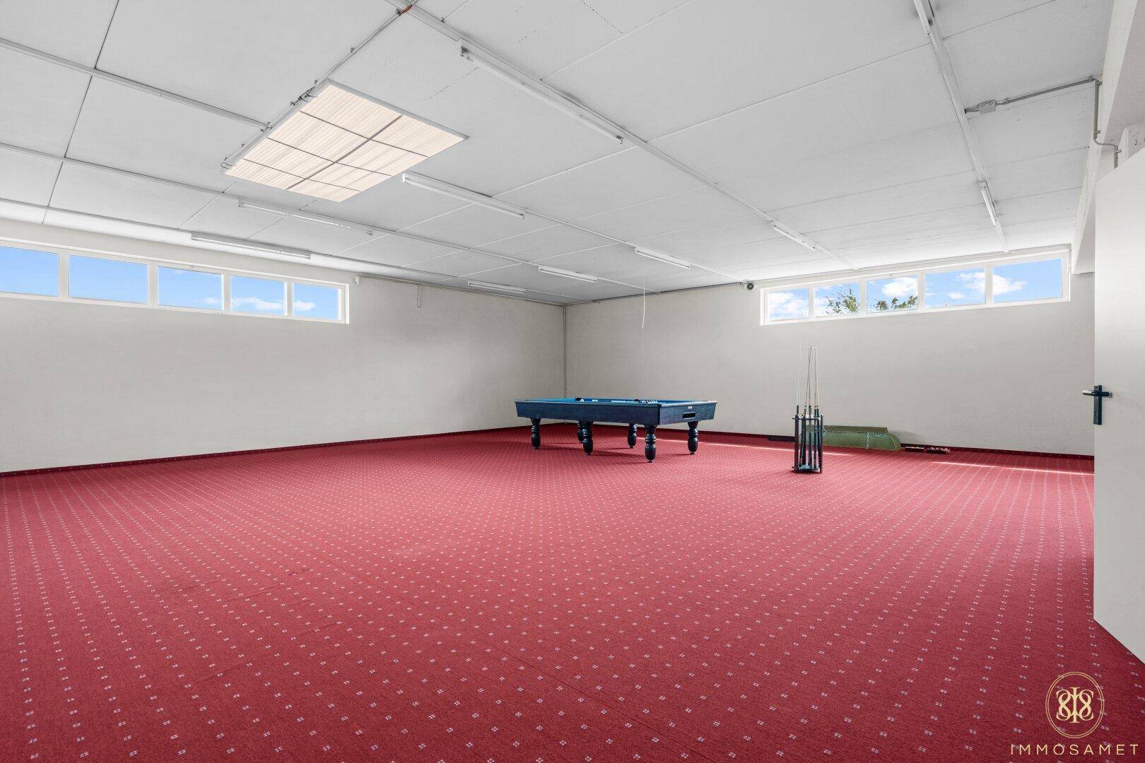 Großer Freizeitraum mit rotem Teppichboden, Billardtisch und hohen Fenstern für Tageslicht.